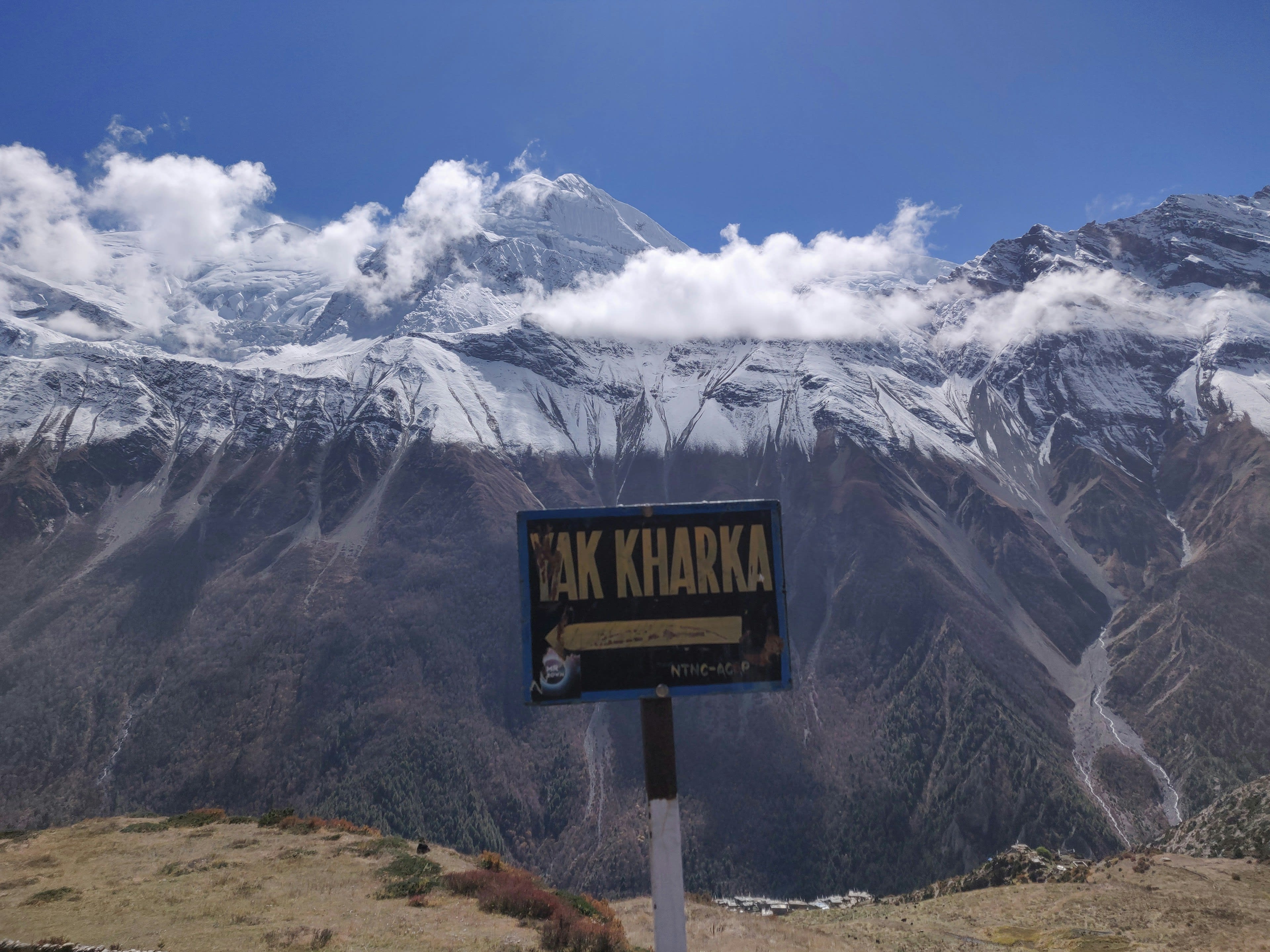 A trail sign at Yak Kharka with dry mountainsides and snow peaks behind