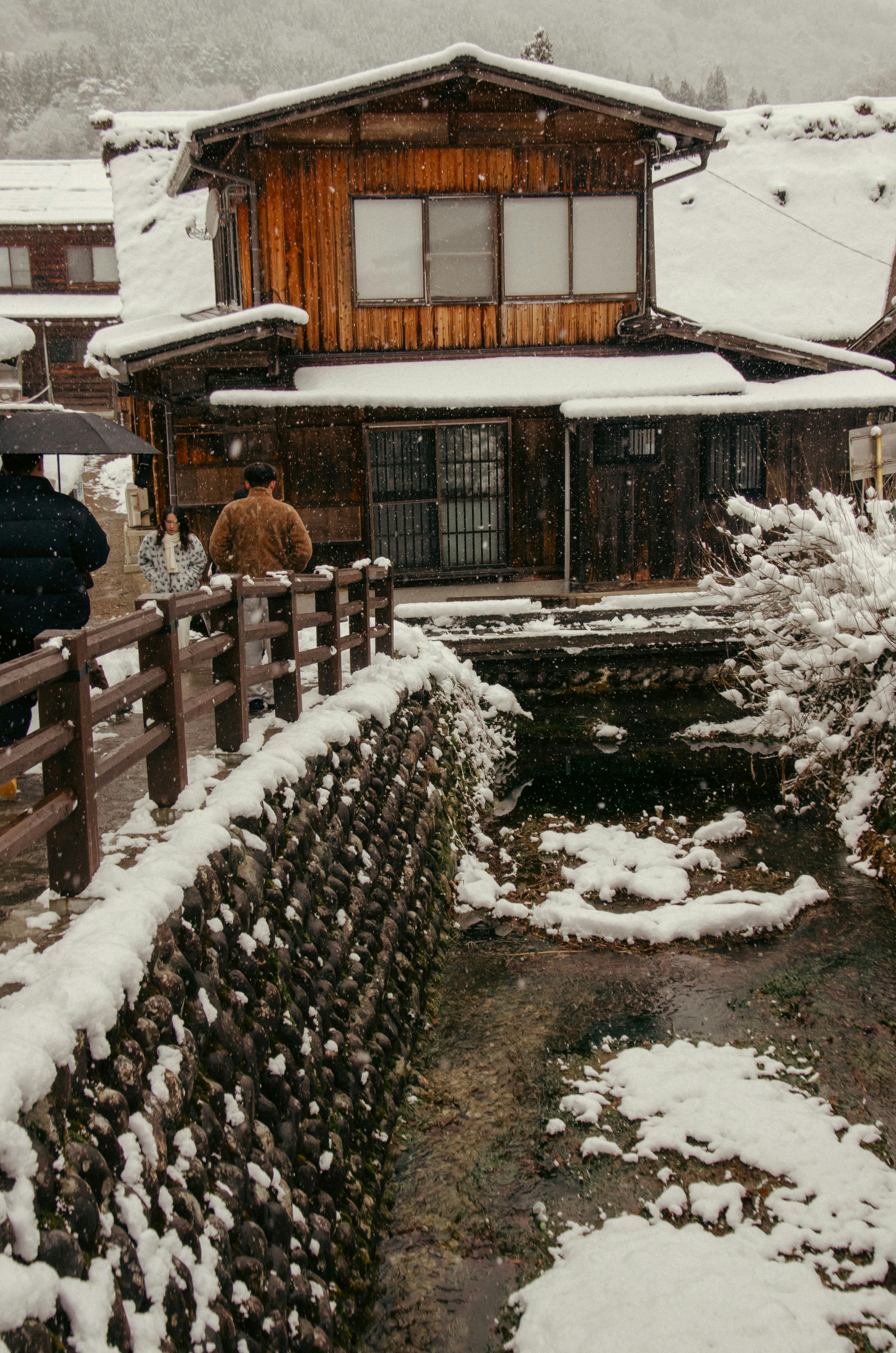 Wooden houses with steep roofs beside a stone-walled stream during heavy snowfall