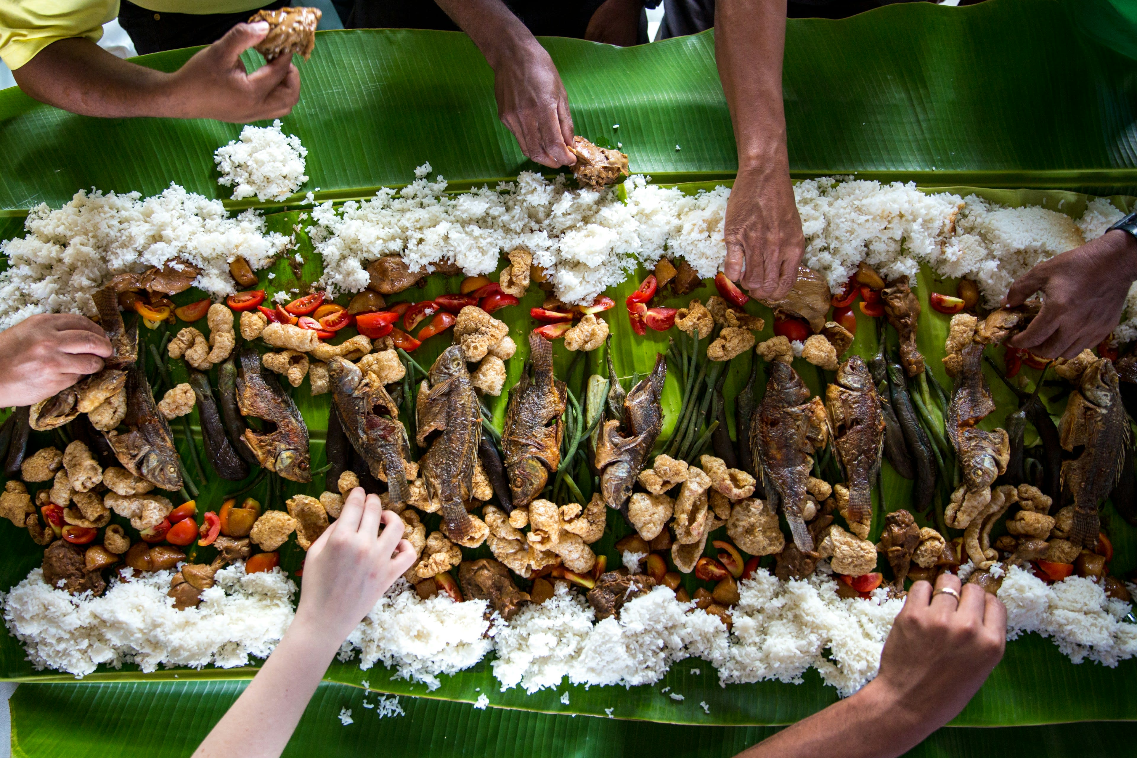 Hands reaching over a banana-leaf spread of grilled fish and rice