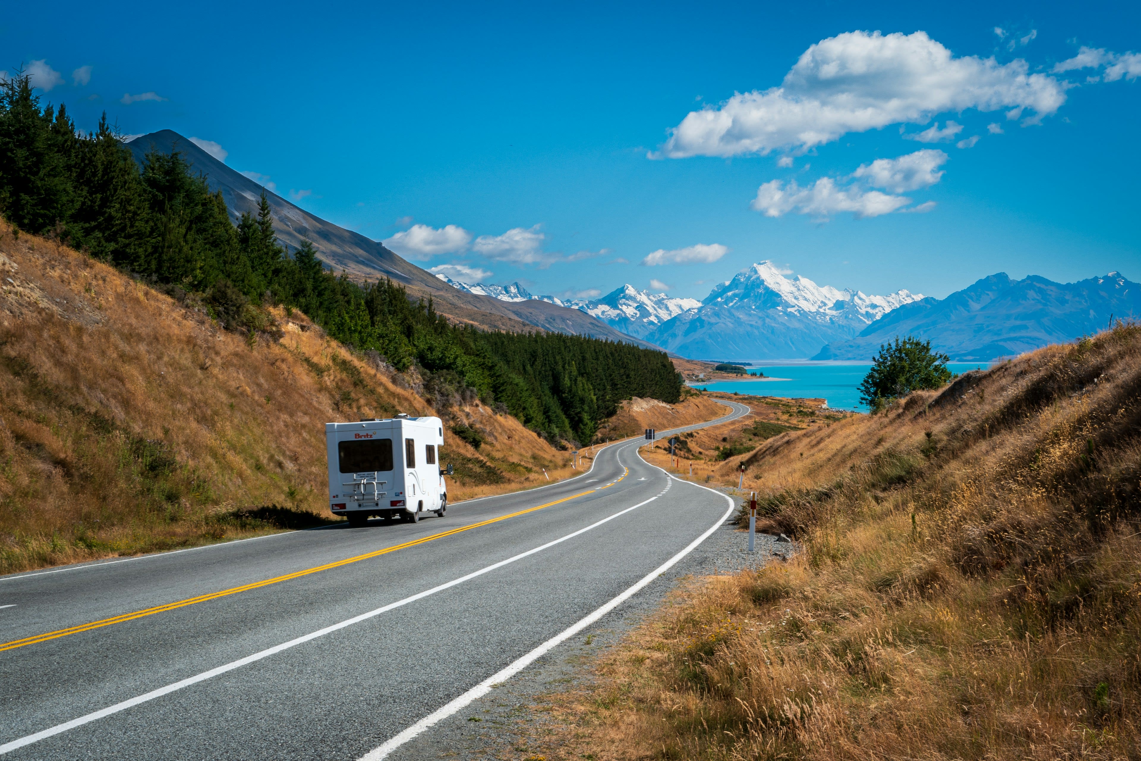 A campervan on a highway curving toward a snow-capped mountain above a turquoise lake
