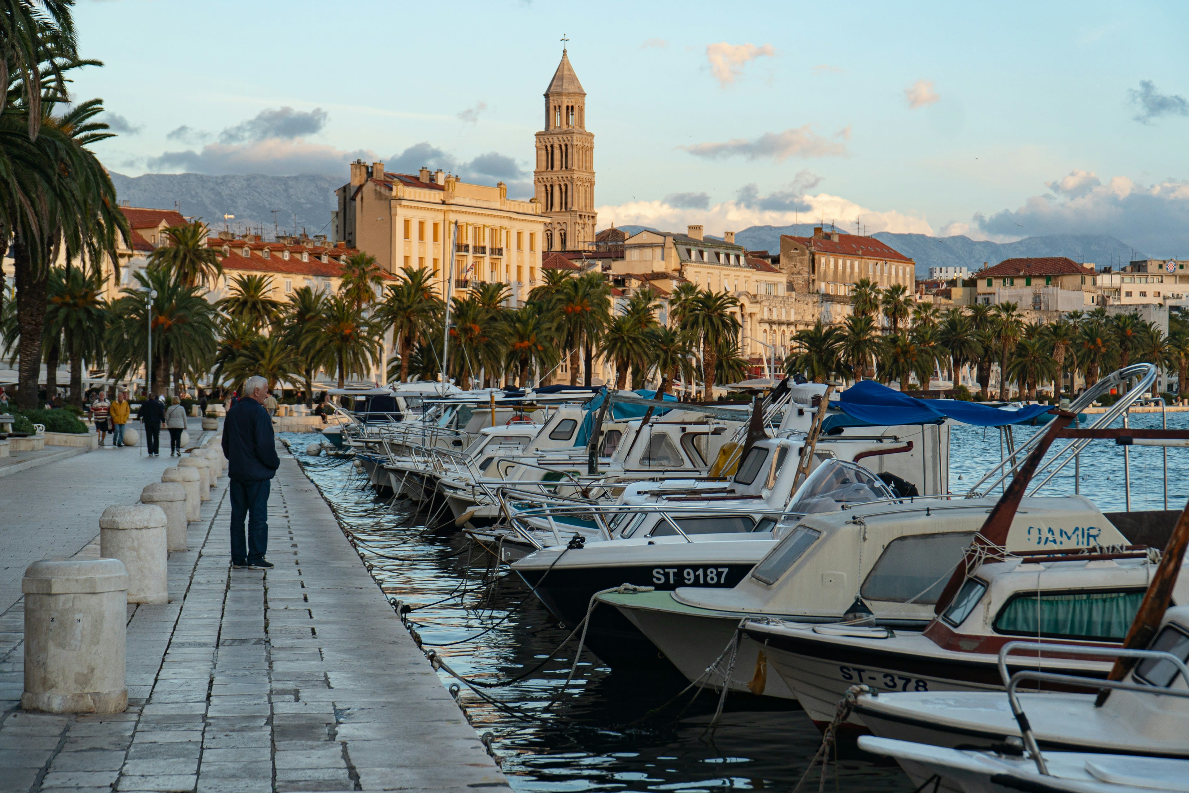 A man walking the palm-lined Riva promenade in Split past moored boats