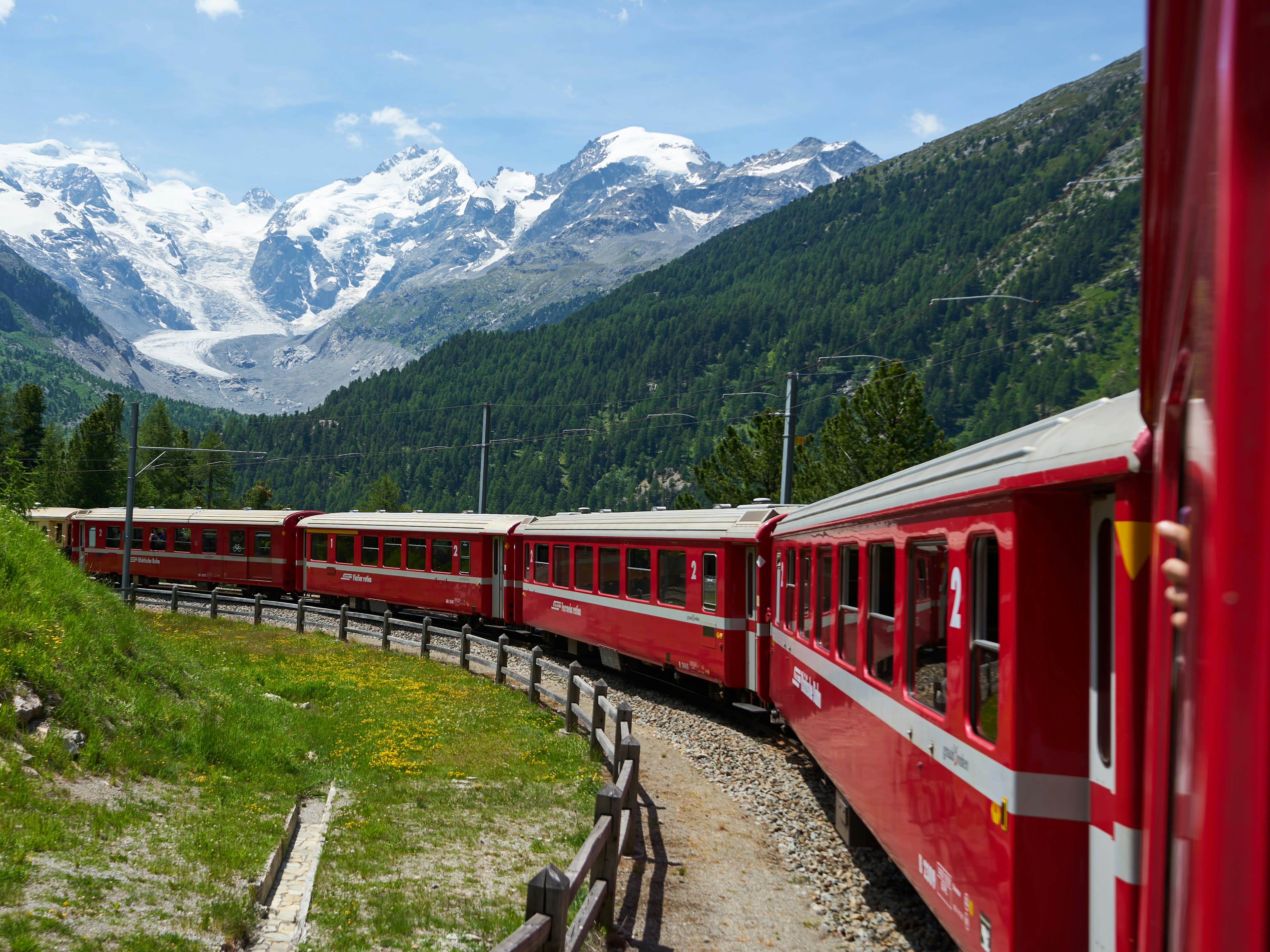 A red train curving through green terrain below high mountains
