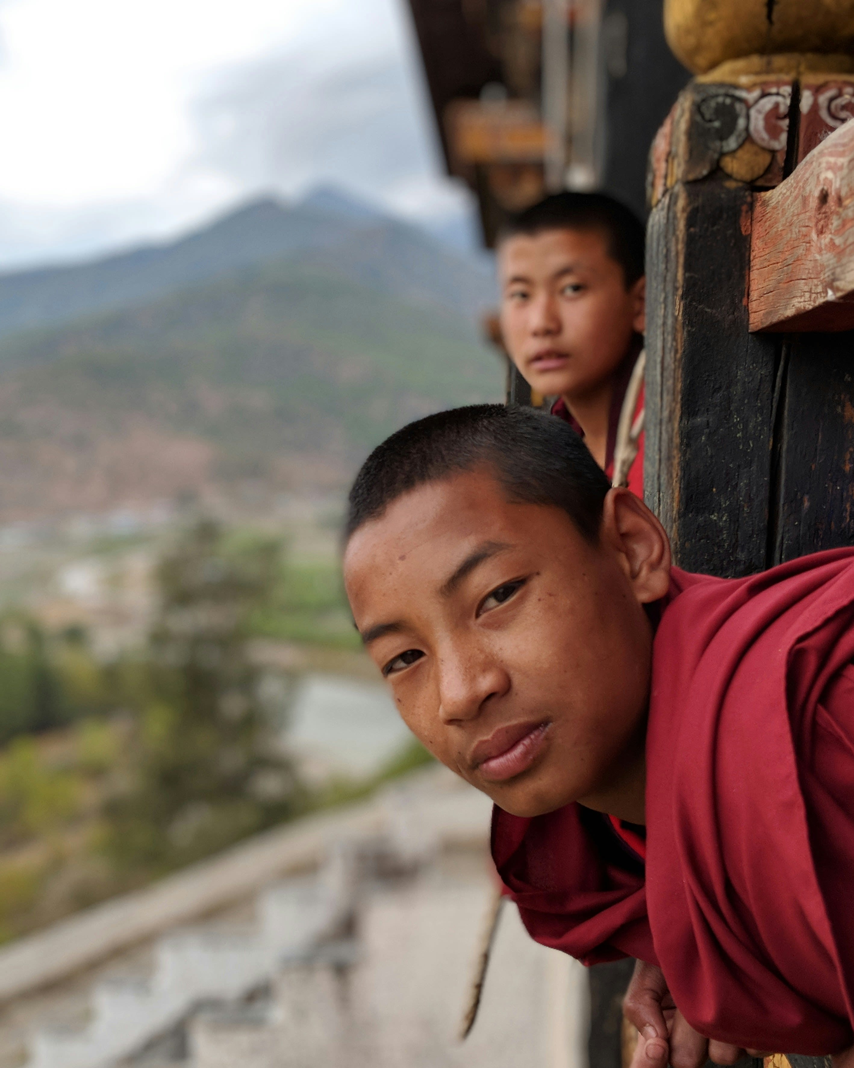 Two young monks in maroon robes looking round a painted wooden pillar