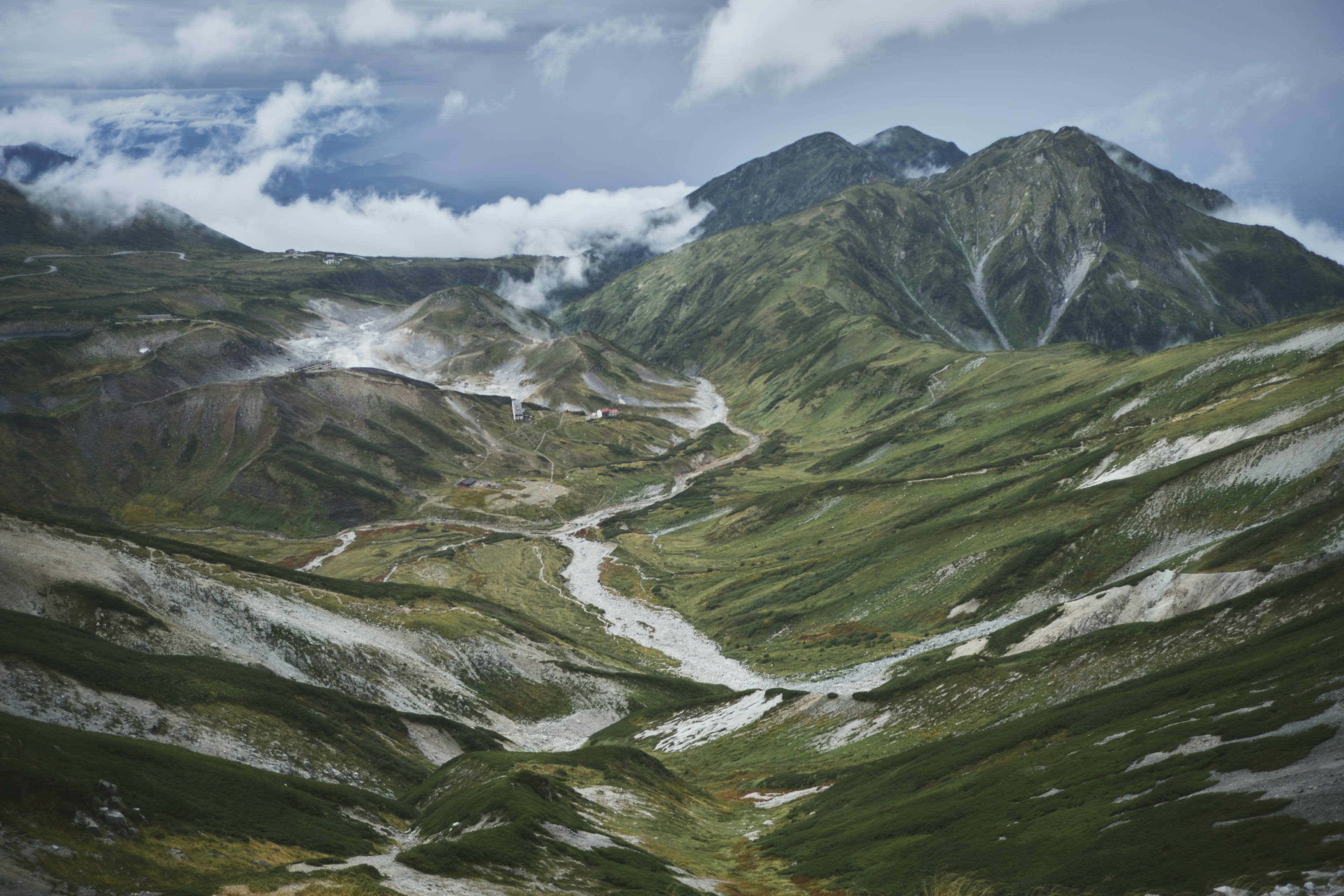 A bare alpine basin of green and grey slopes streaked with old snow under cloud