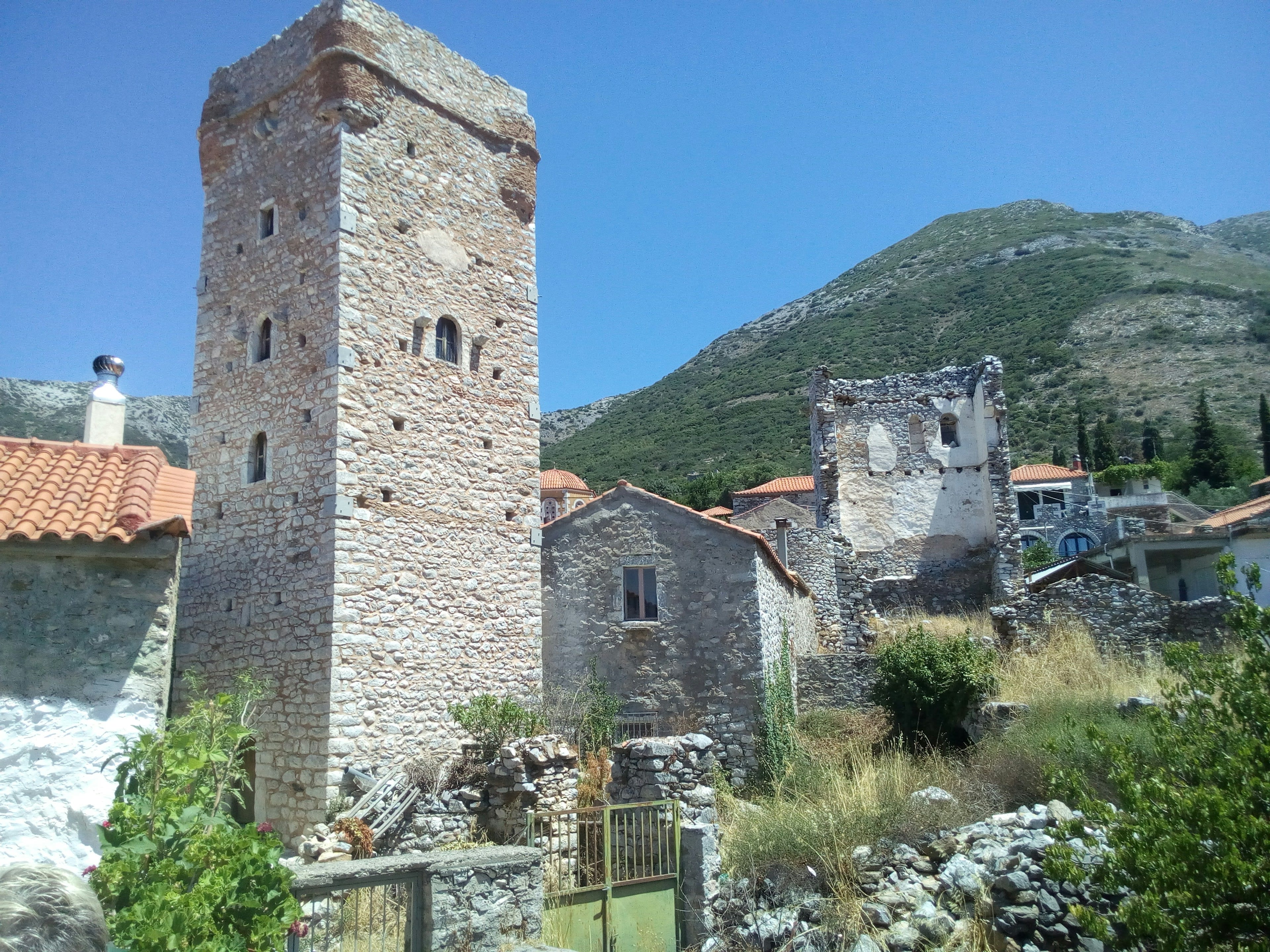 A tall dry-stone defensive tower beside older stone buildings with a green hill behind