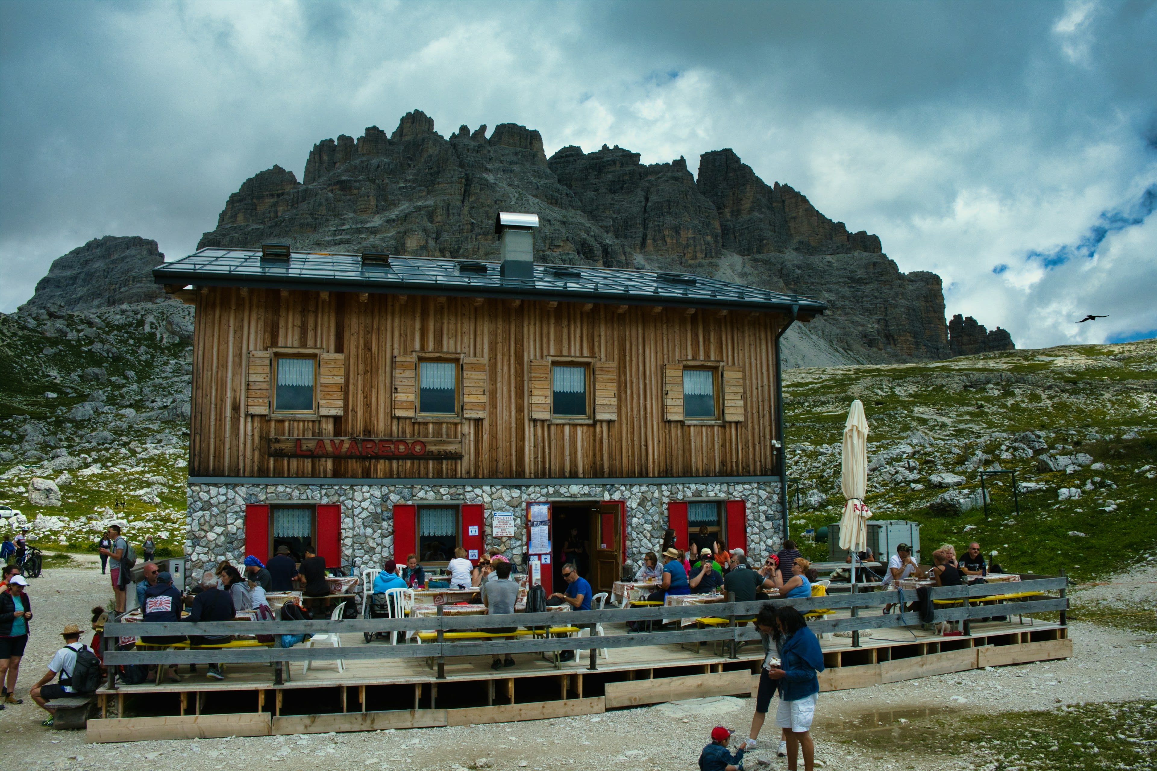 A wooden mountain lodge with hikers crowded onto its patio under grey peaks