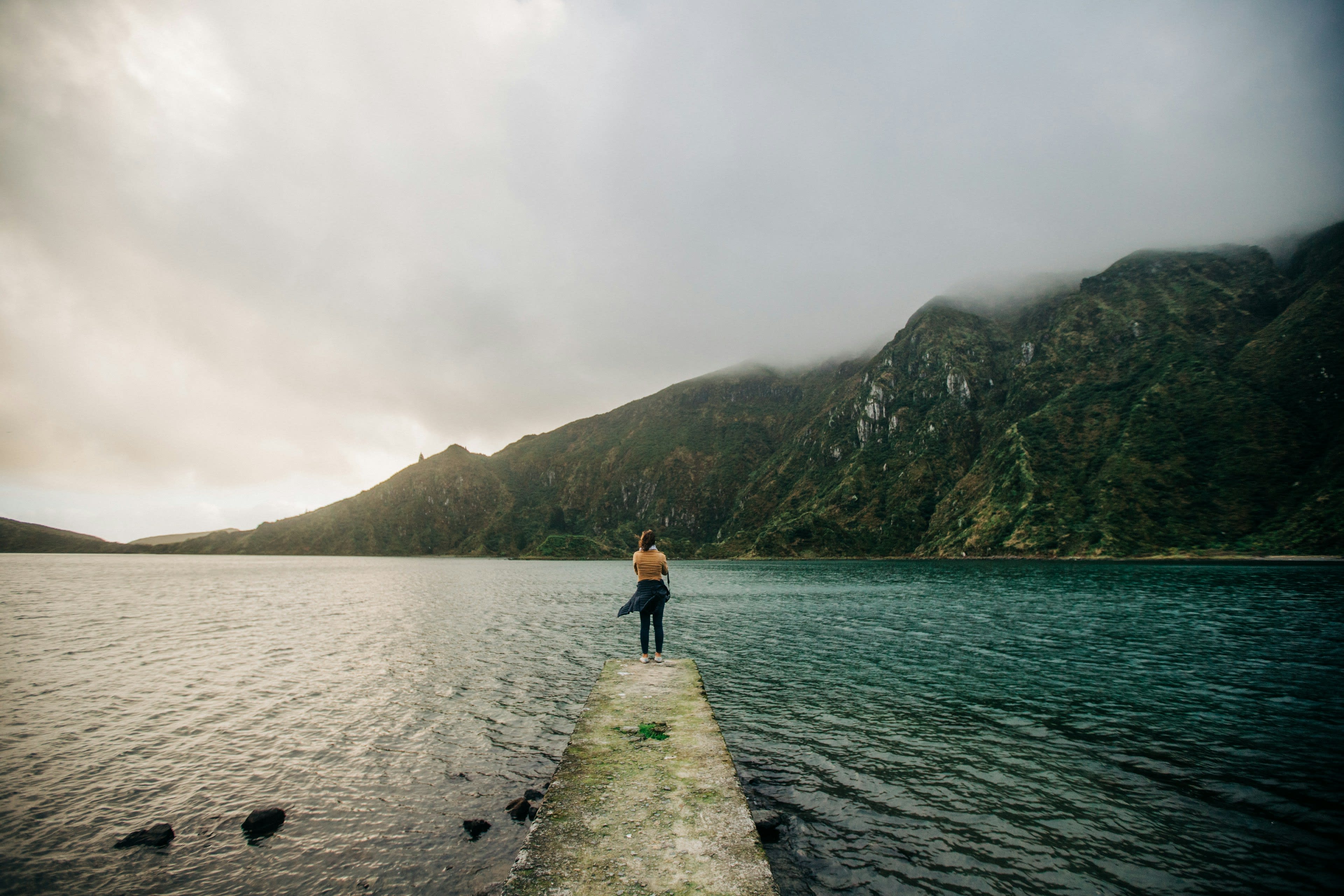 A person standing at the end of a jetty over a still lake with mist on the far shore