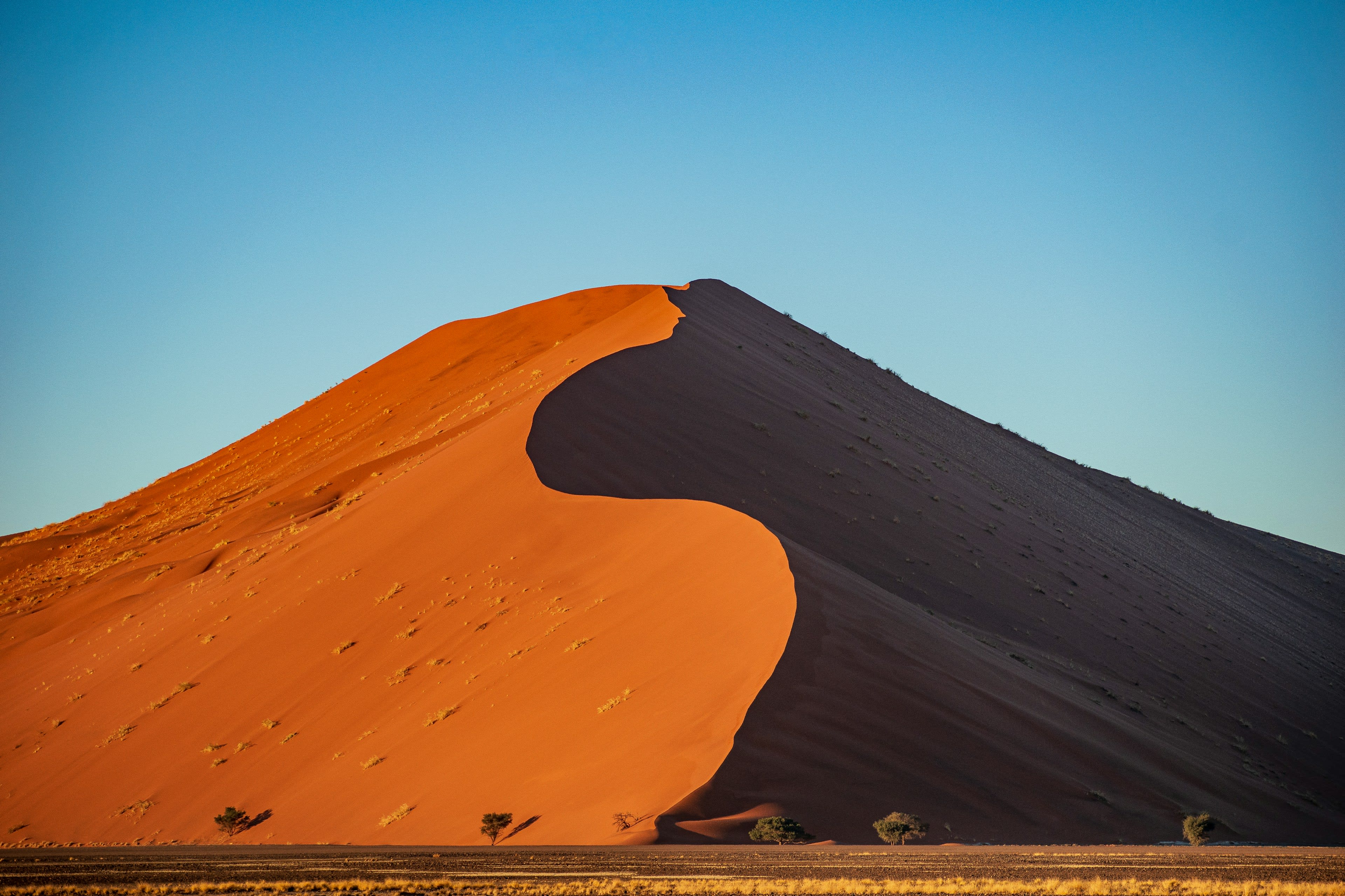 A large orange sand dune with one lit face and one in deep shadow under blue sky