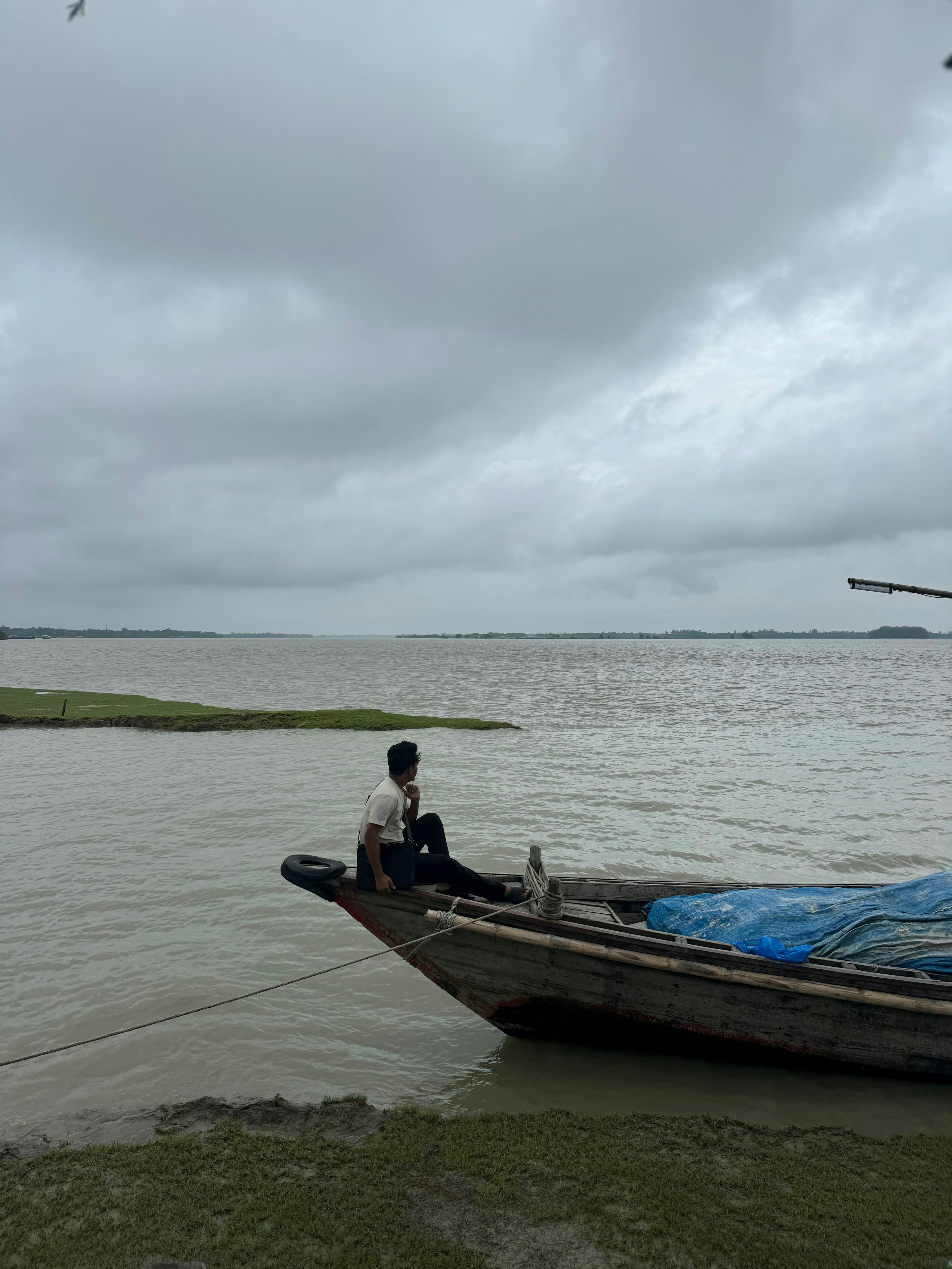 A man sitting on the side of a wooden boat on a wide grey river under heavy cloud