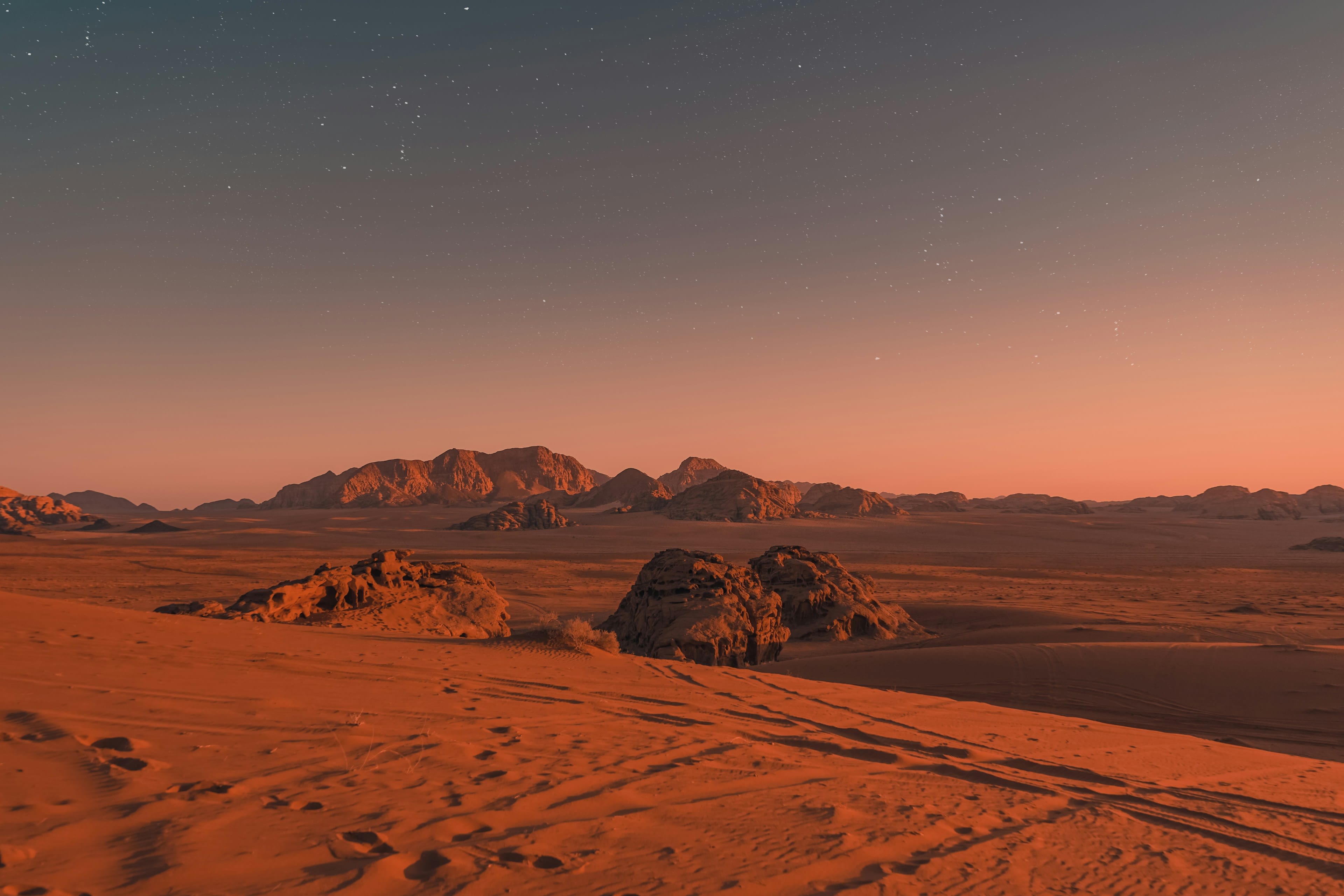 A flat red sand plain with tyre tracks and eroded sandstone mountains behind