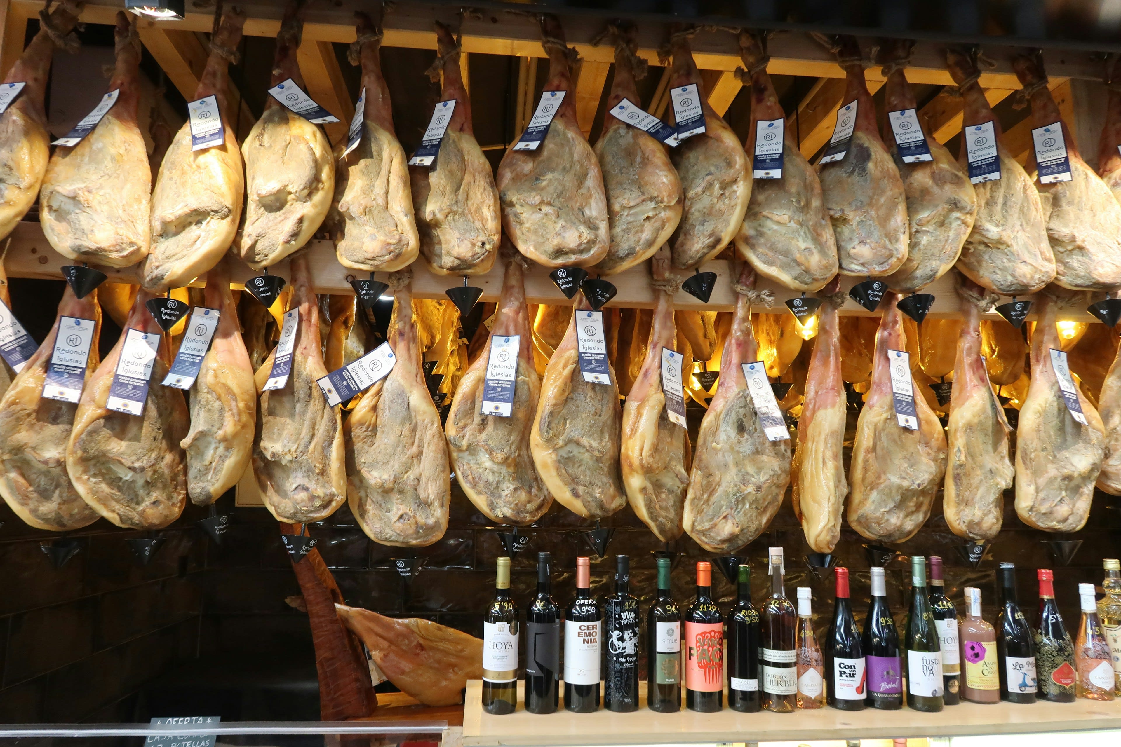 Rows of cured ham legs hanging above shelves of wine in a shop