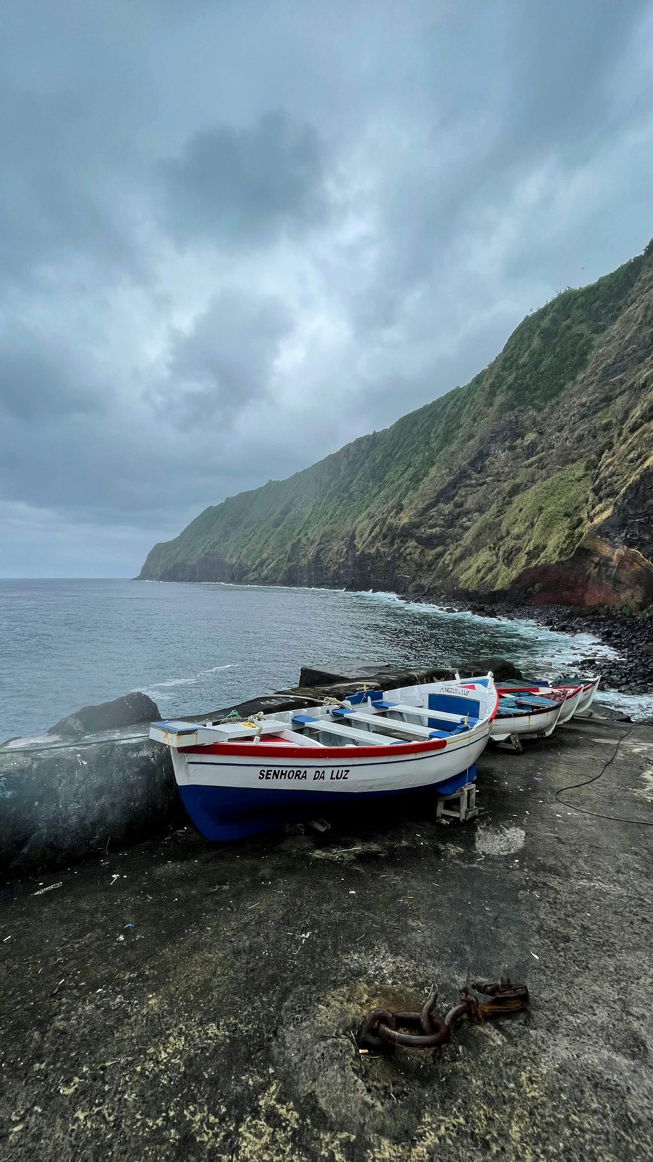 A small wooden boat on a stony shore below a huge green sea cliff