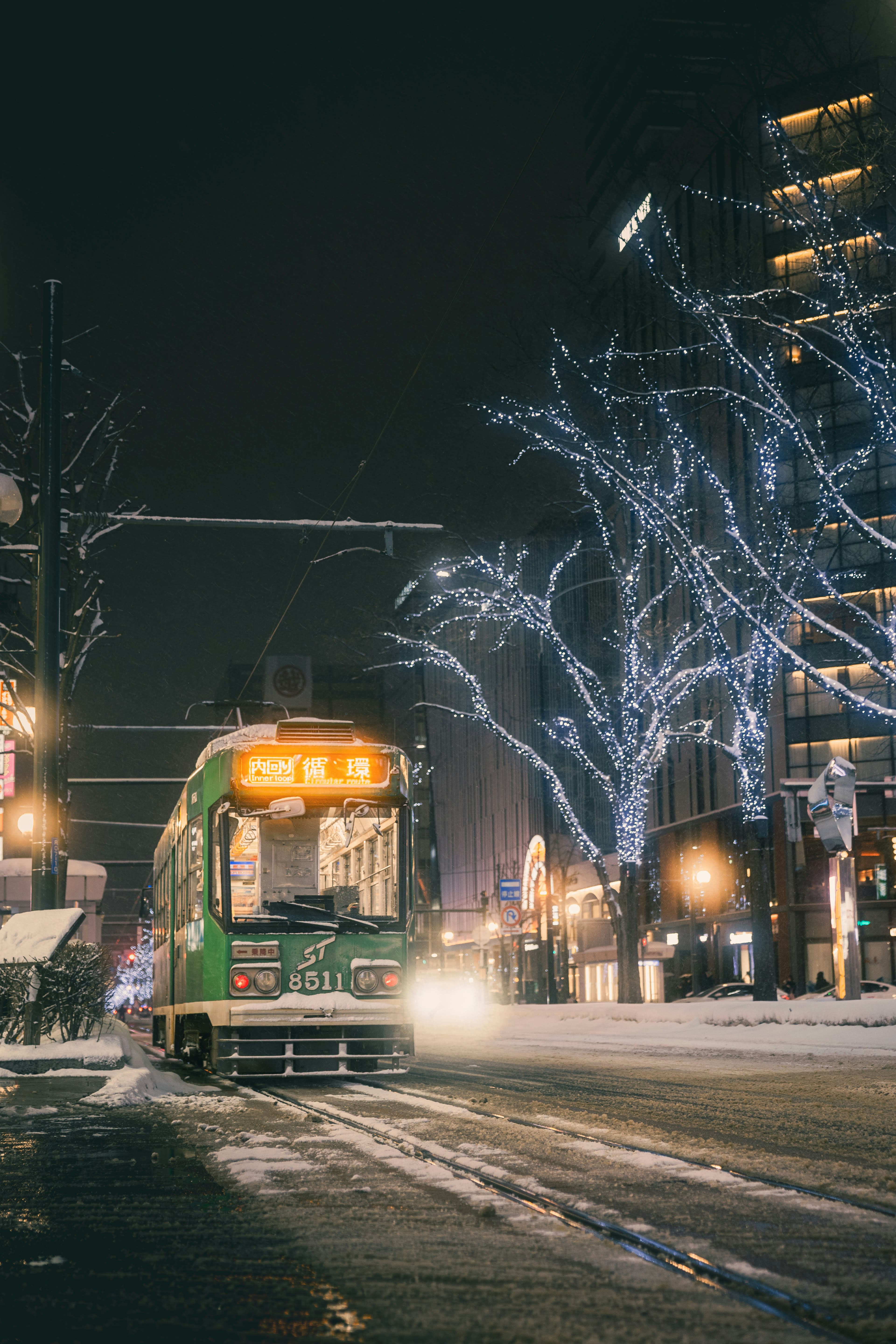 A green tram moving through falling snow on a Sapporo street at night