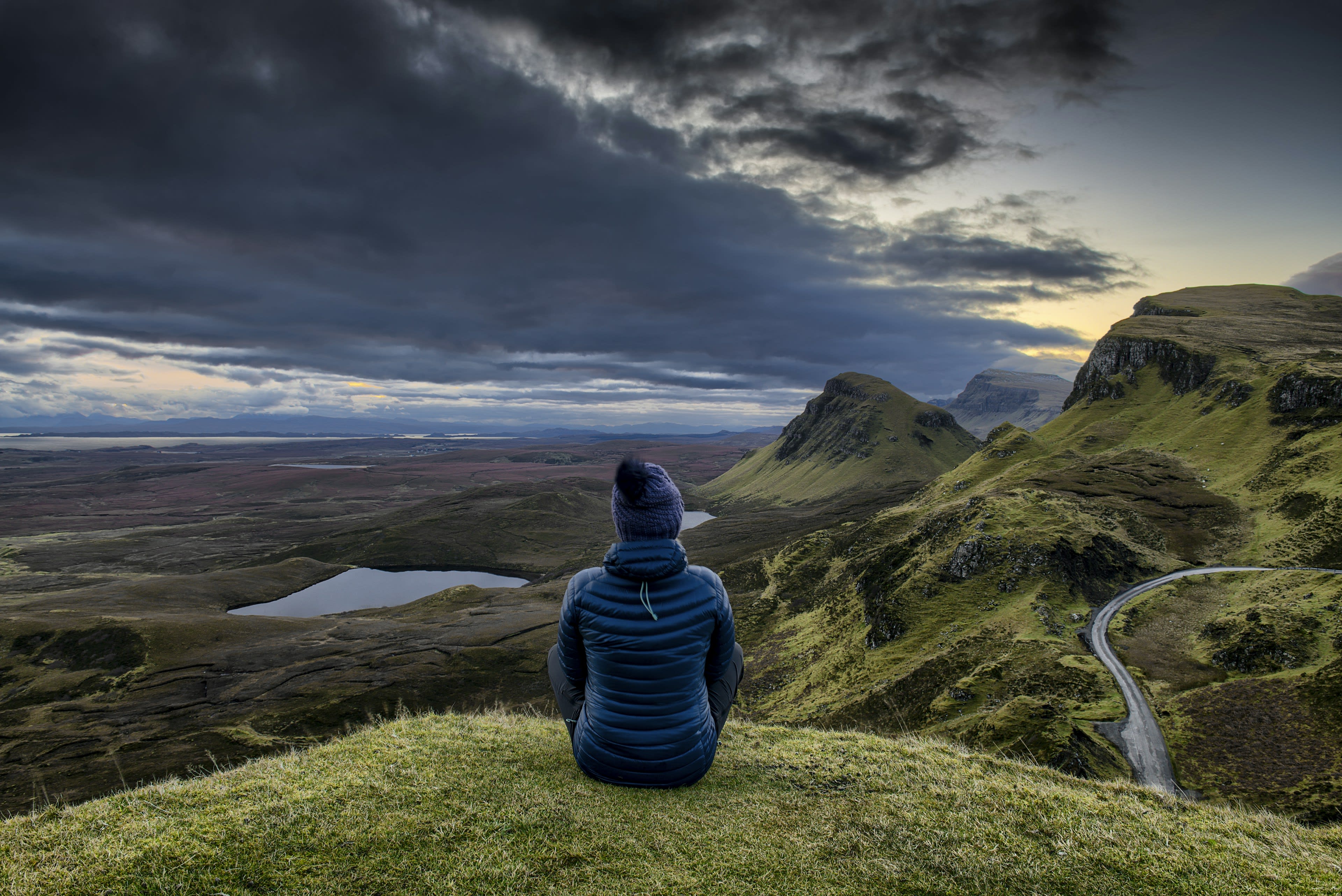 A person in a blue hooded coat sitting on grass above a wide green landslip landscape