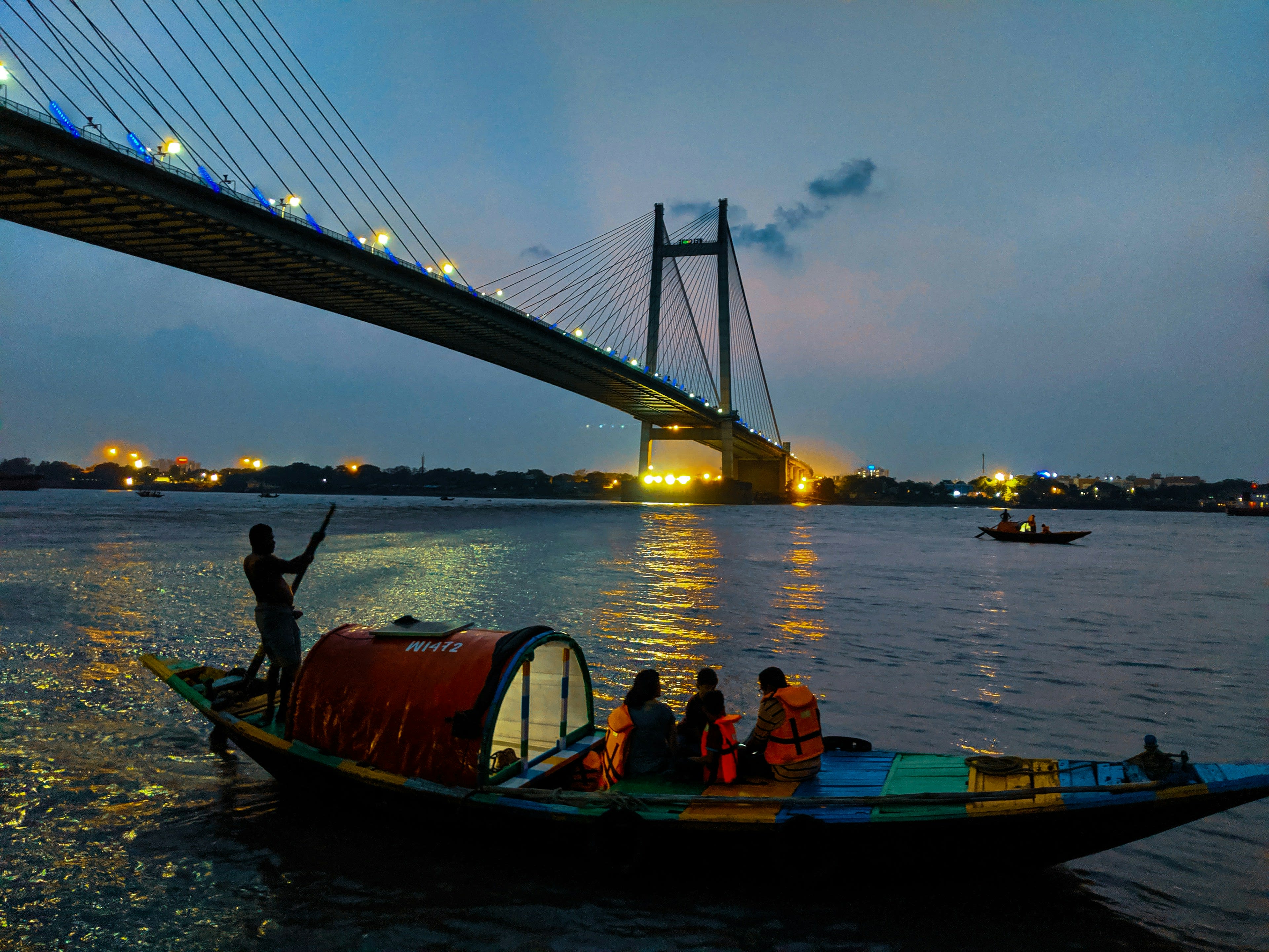 A crowded country boat crossing a river at dusk beneath a lit cable-stayed bridge