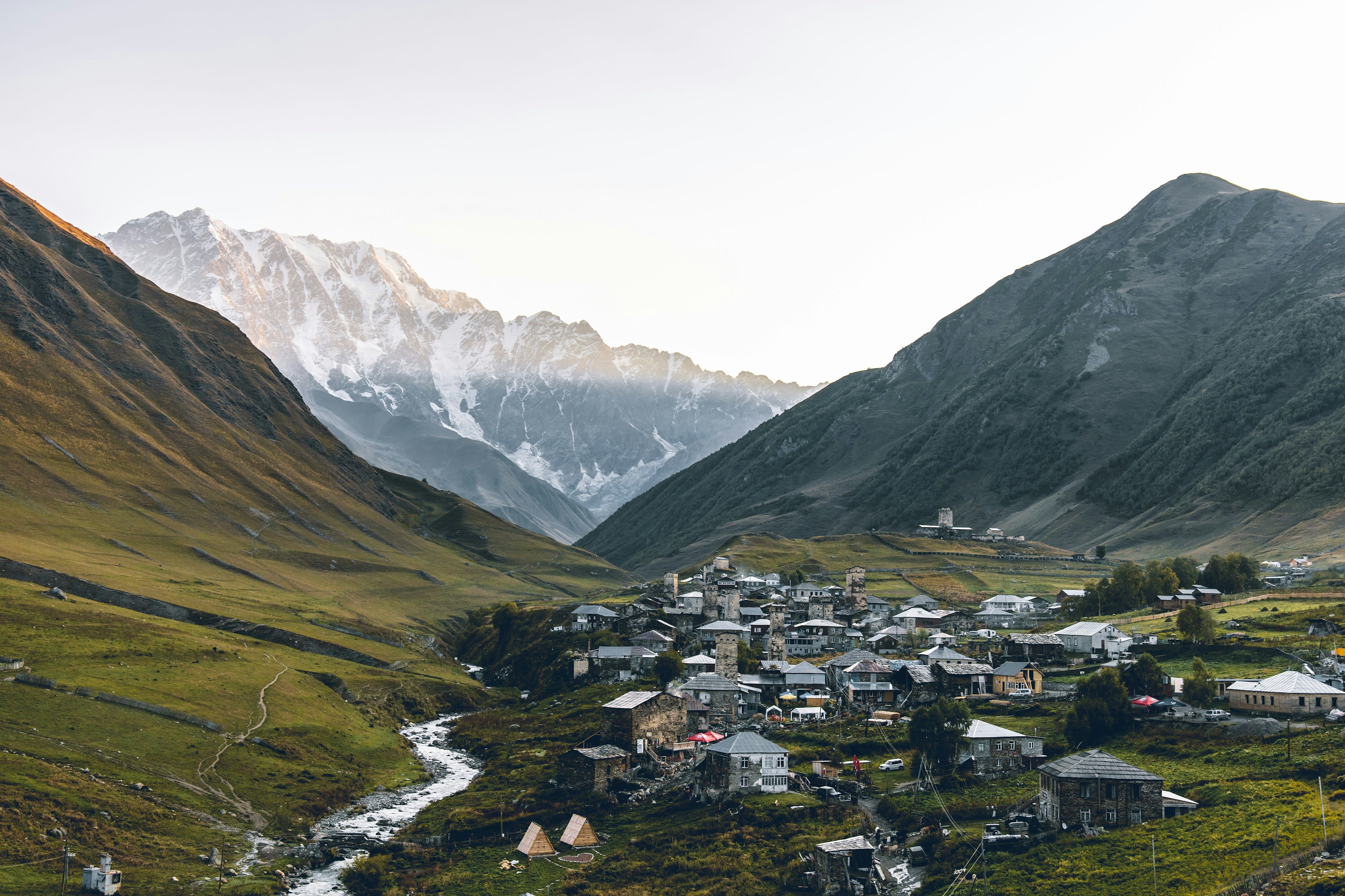 A stone village in a wide green valley seen from above with snow peaks beyond