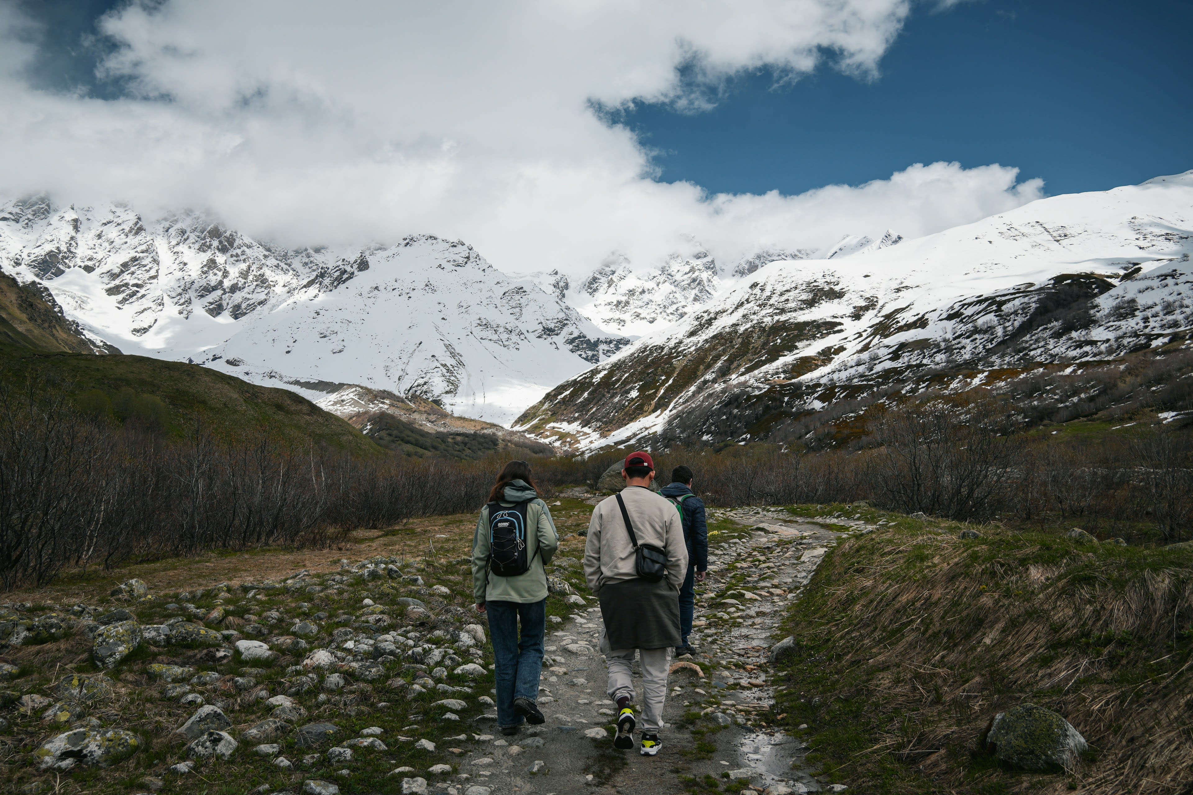 Two walkers on a dirt track heading towards a wide snow-covered mountain range