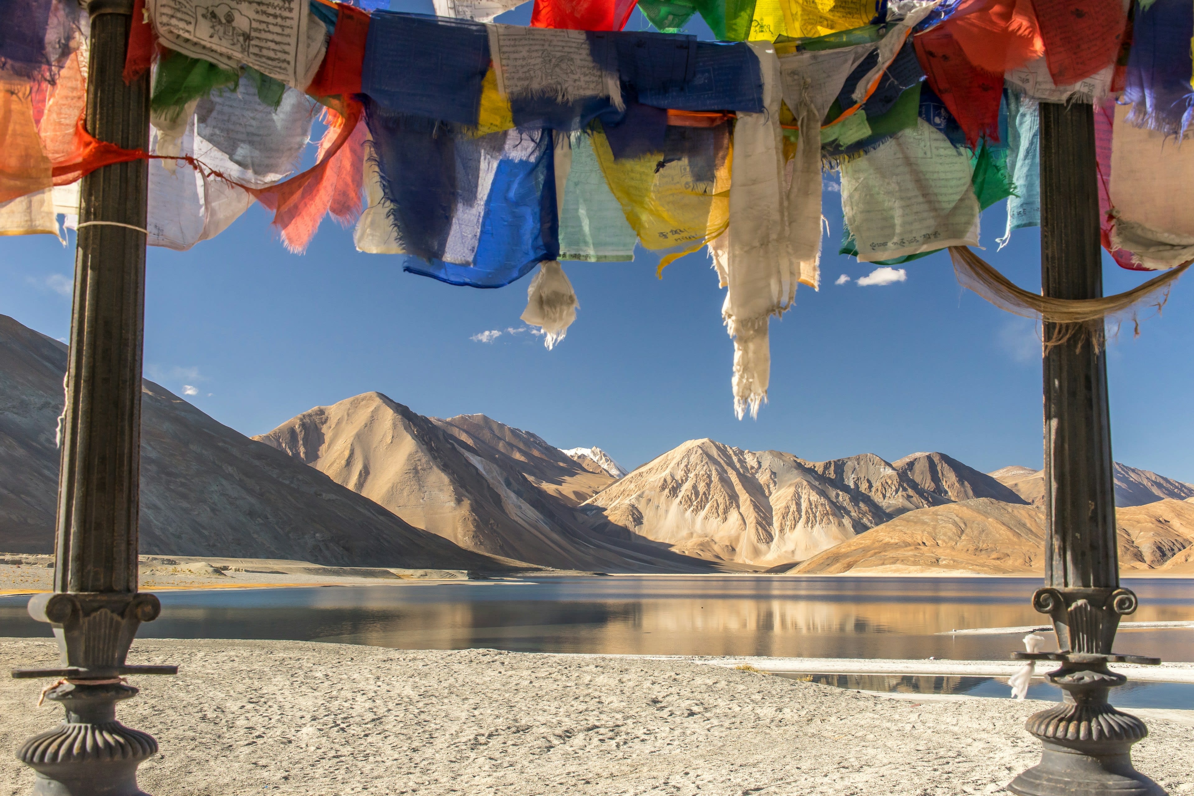 Prayer flags strung above a bright blue high-altitude lake with brown mountains beyond