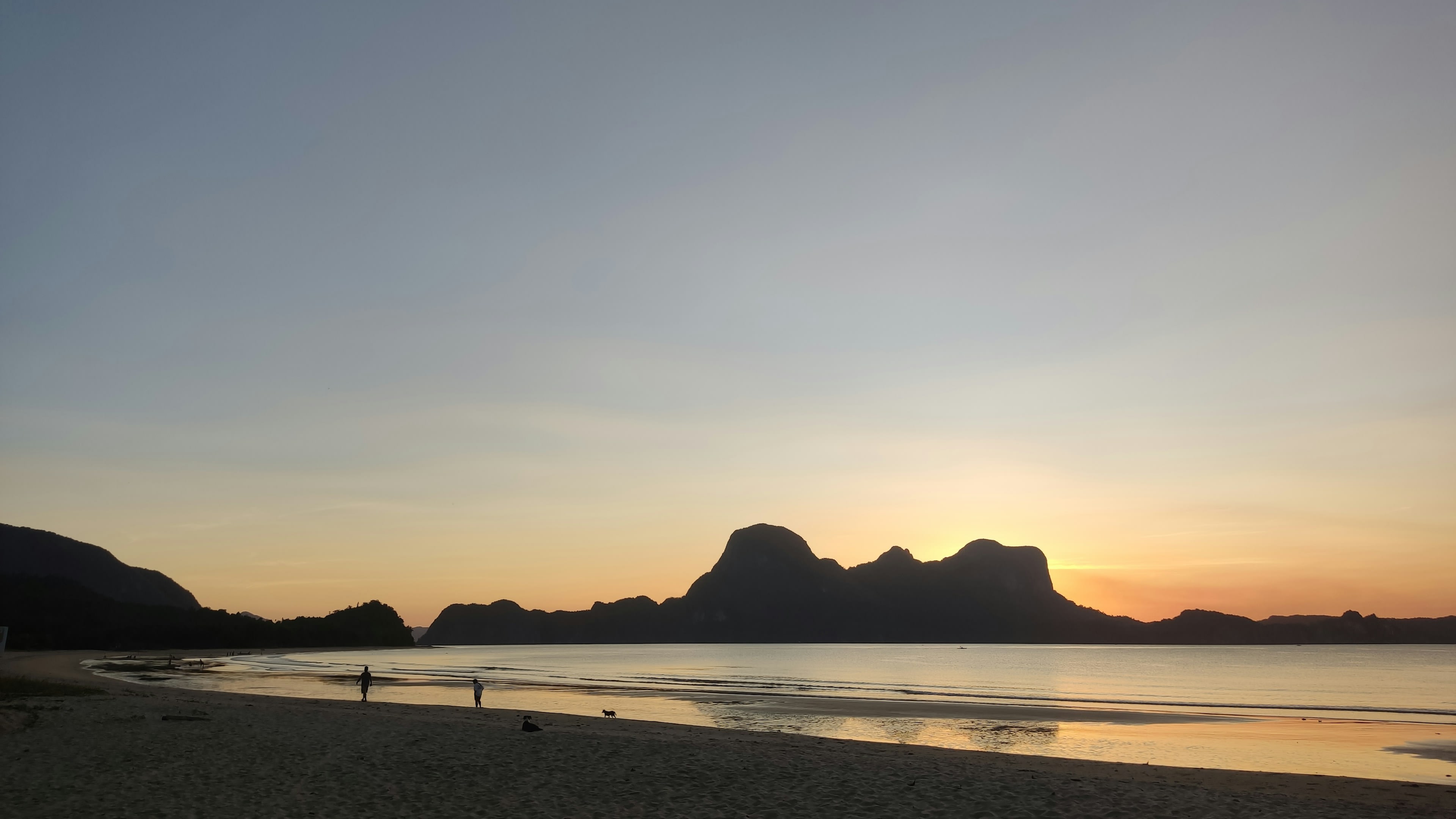 People on a beach at sunset with hills and boats in silhouette