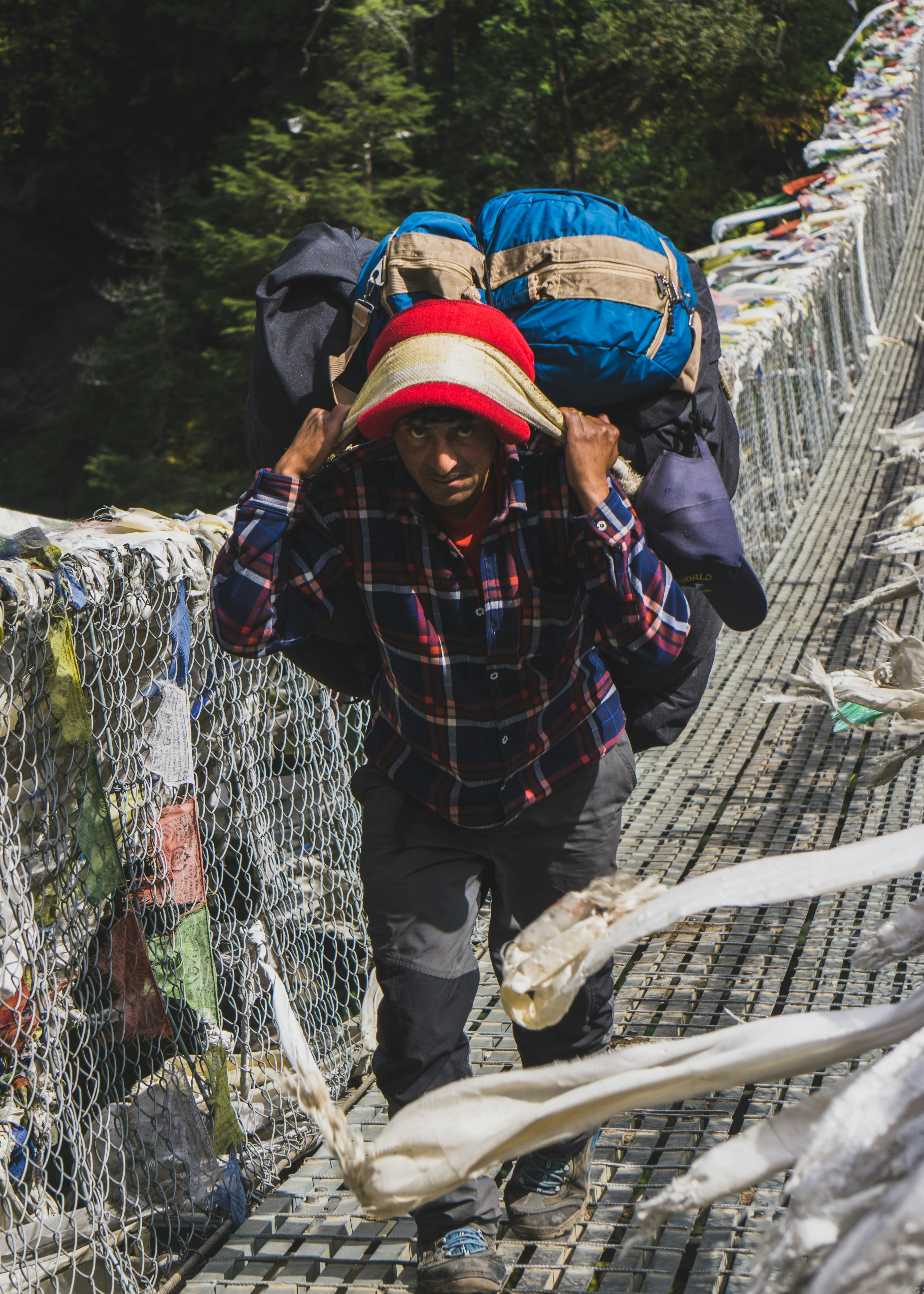 A porter carrying a heavy load across a suspension bridge strung with prayer flags