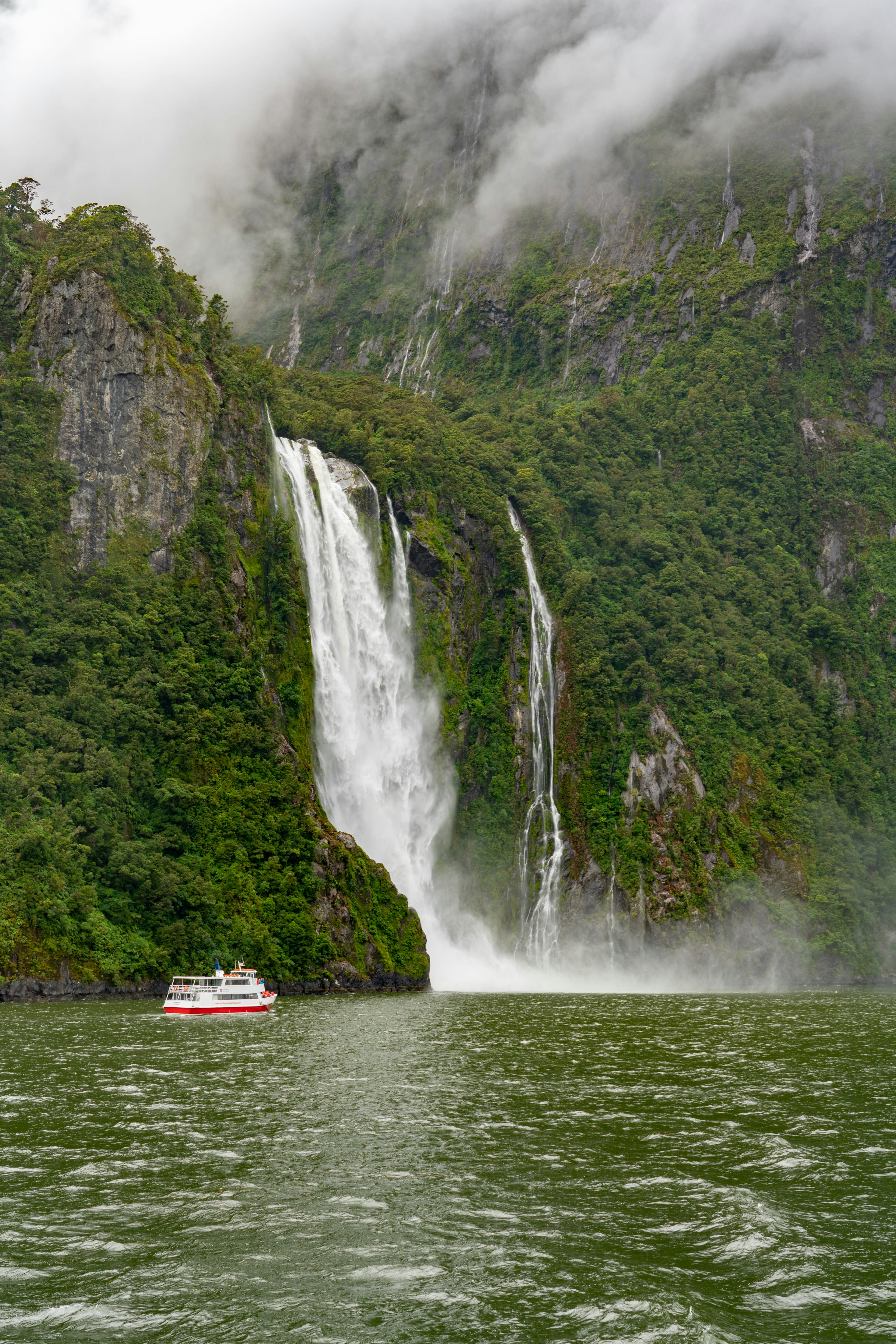A waterfall pouring off a cliff onto a boat in a fjord