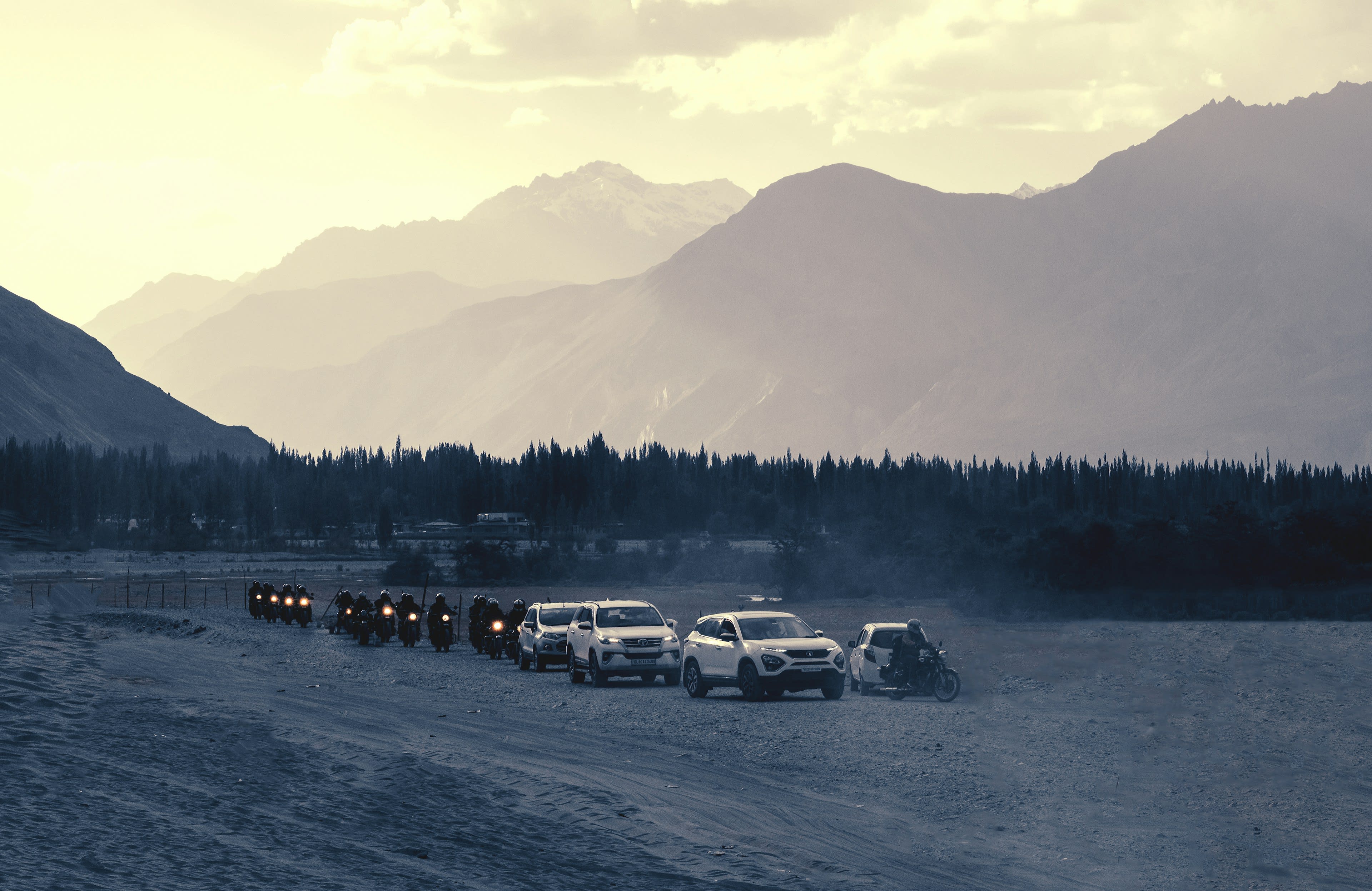 A line of vehicles on a dusty plain beneath snow-streaked mountains