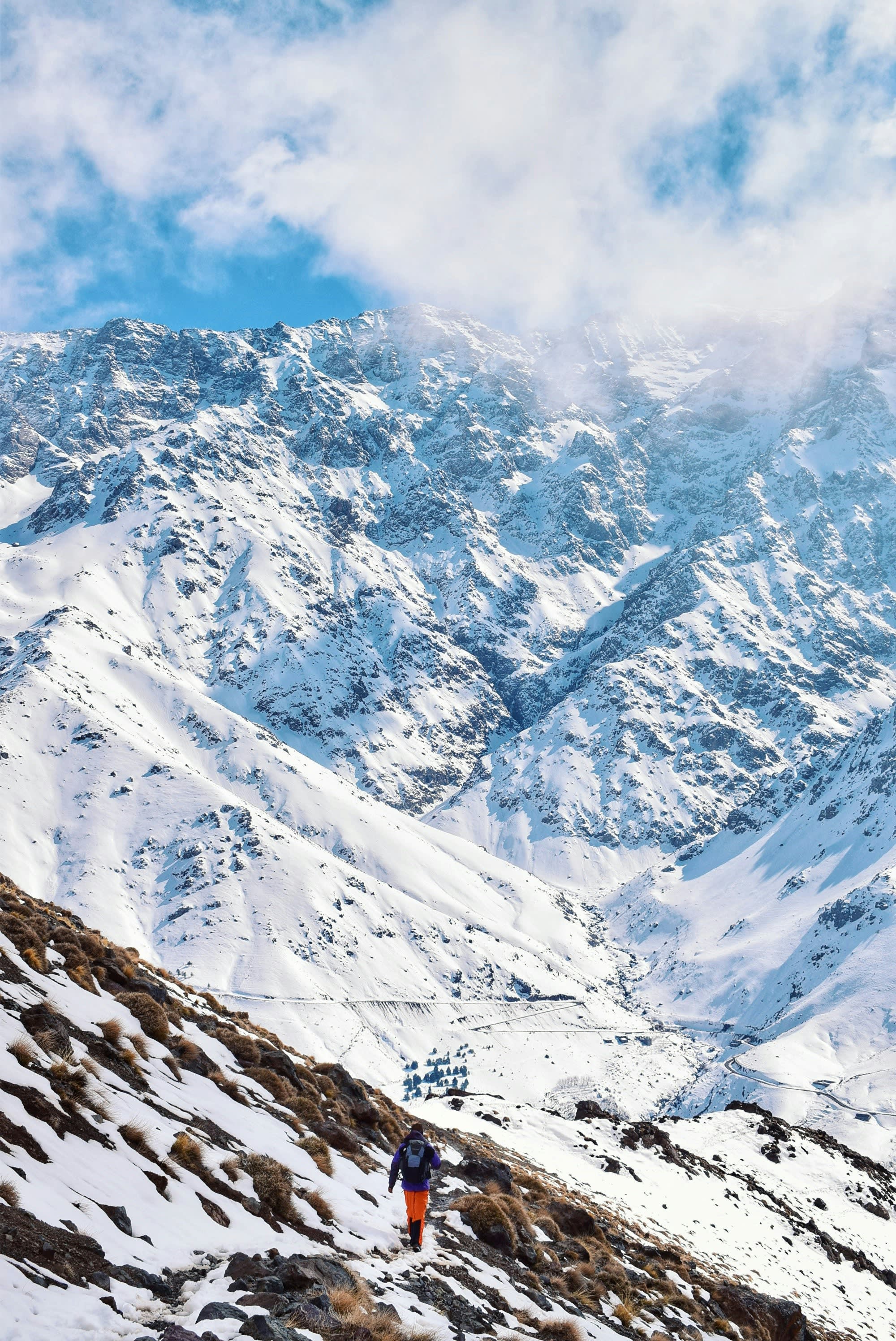 A snow-covered mountain slope with a single tiny figure in red crossing it
