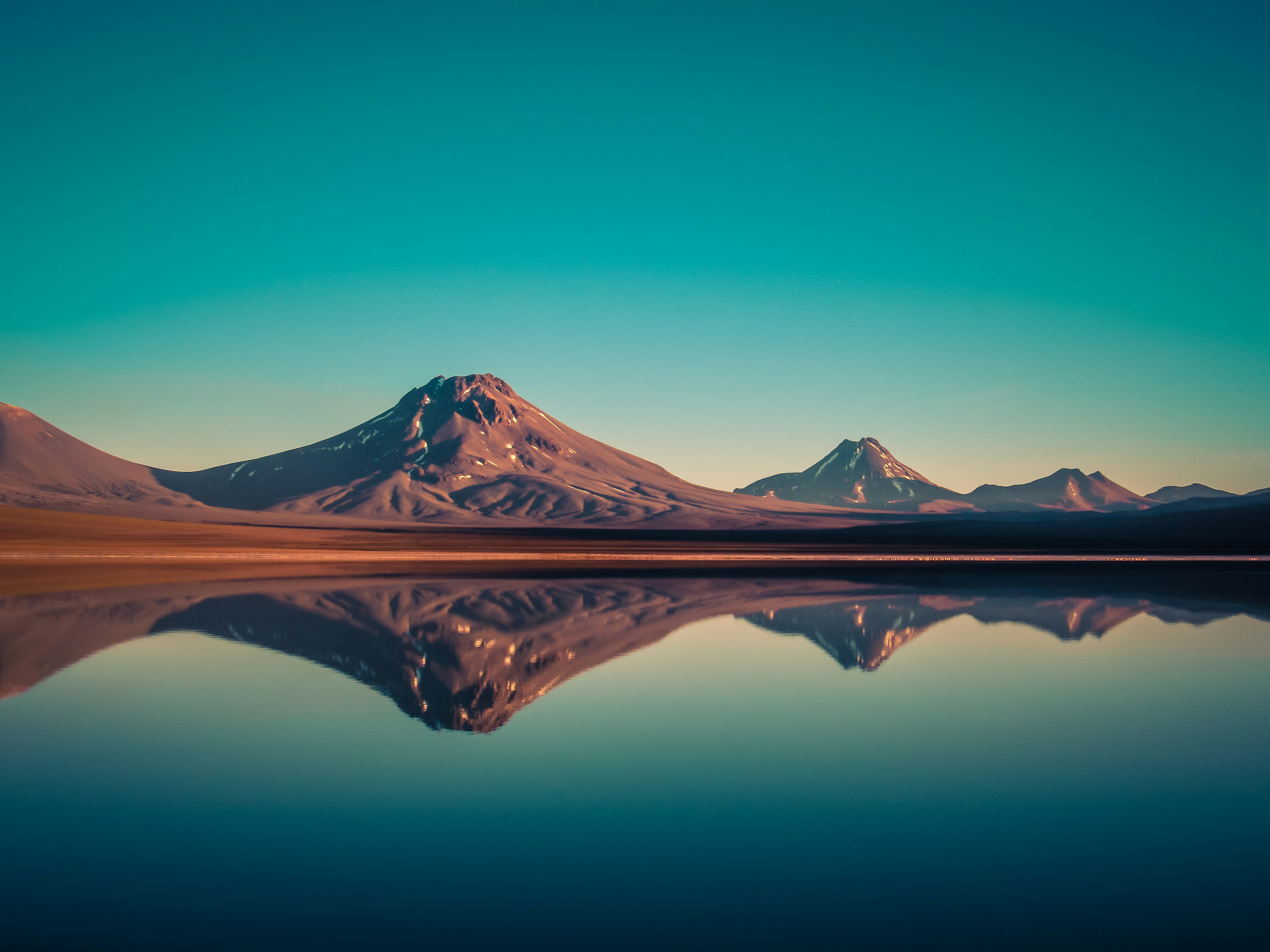 A volcano and its perfect reflection in a still lagoon at dawn, sky shading from teal to rose
