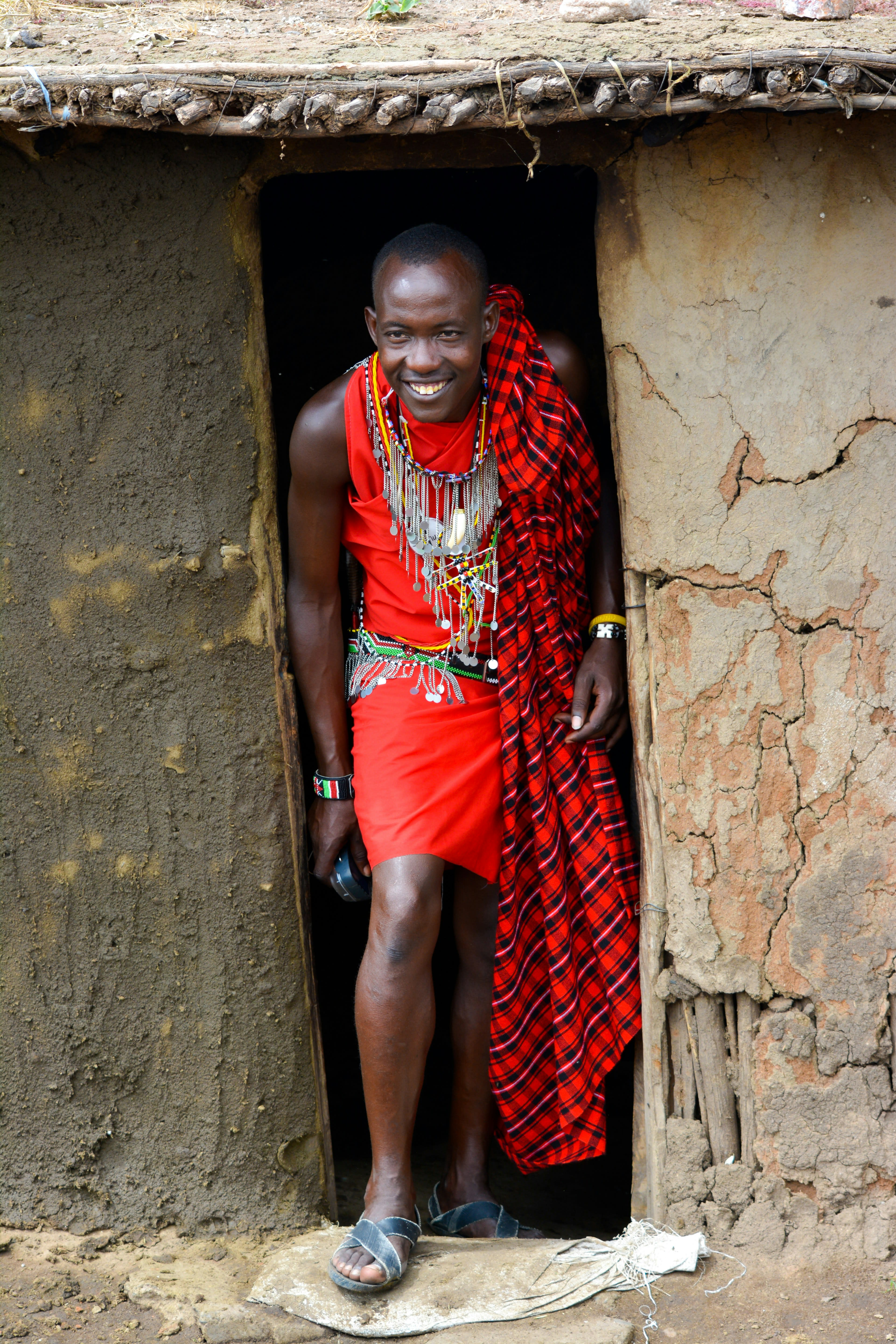 A man in a red shuka standing in the low doorway of a mud-walled house