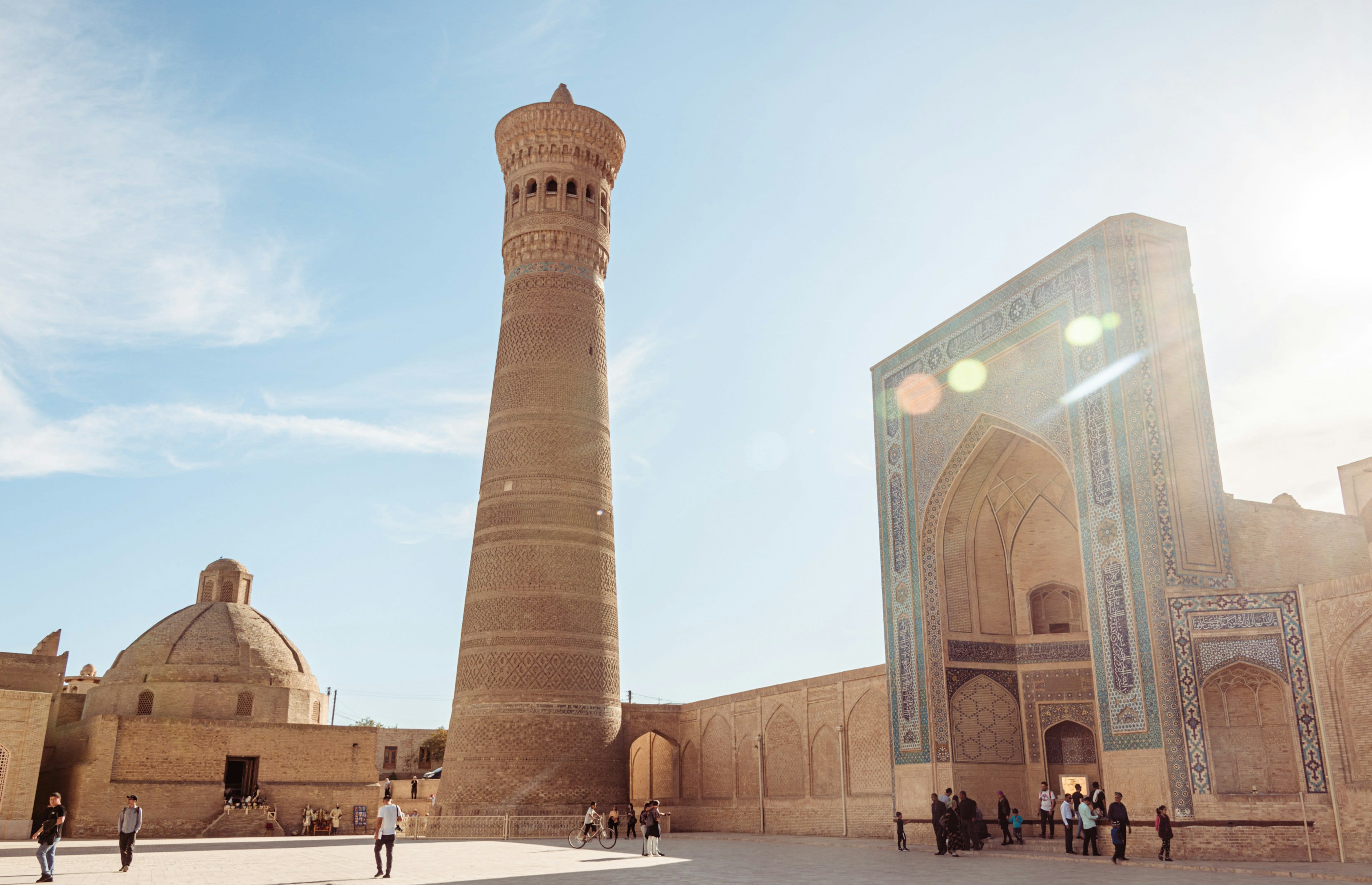 A tall patterned brick minaret beside a tiled mosque portal against the sun