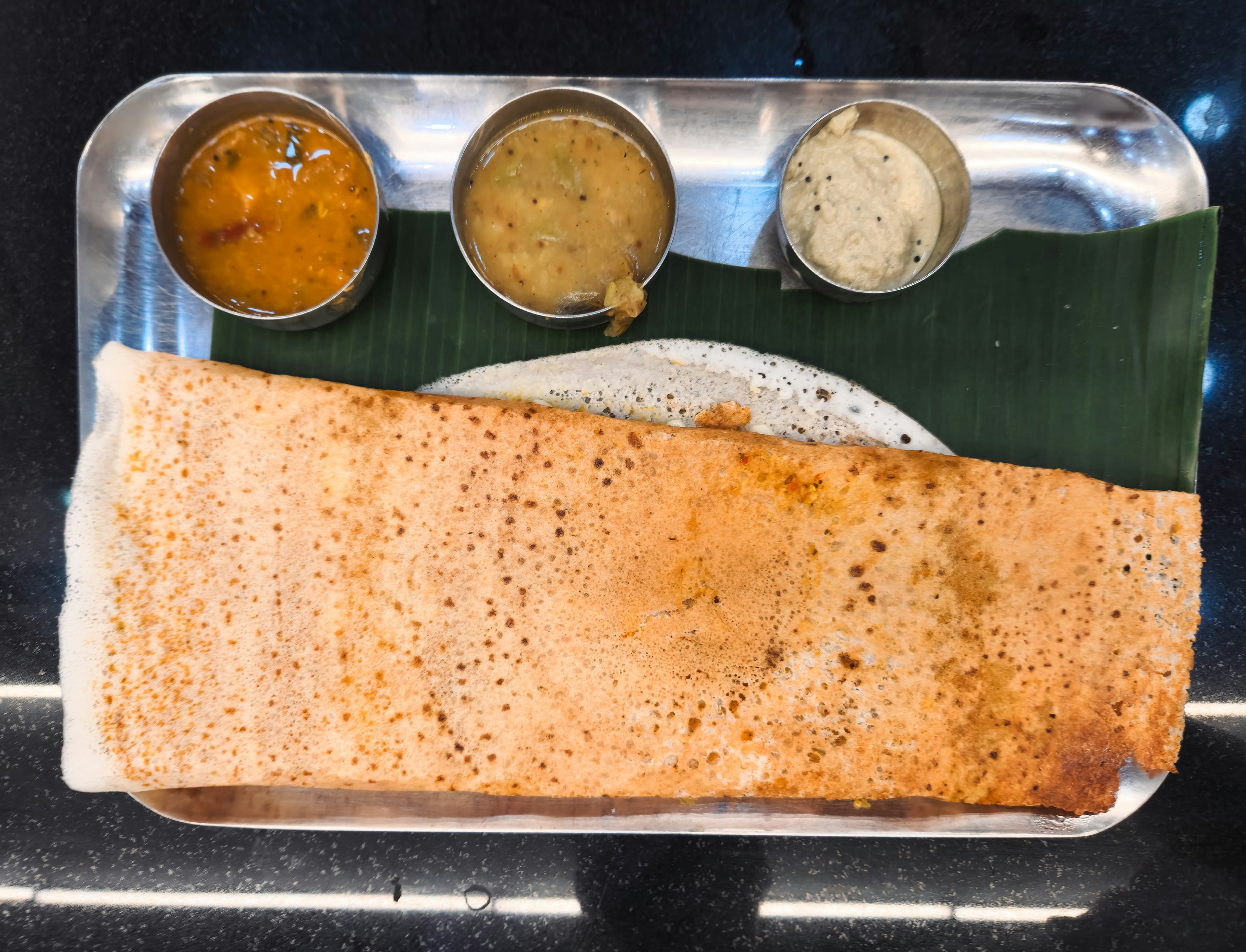 A folded dosa on a steel tray with small bowls of chutney and sambar