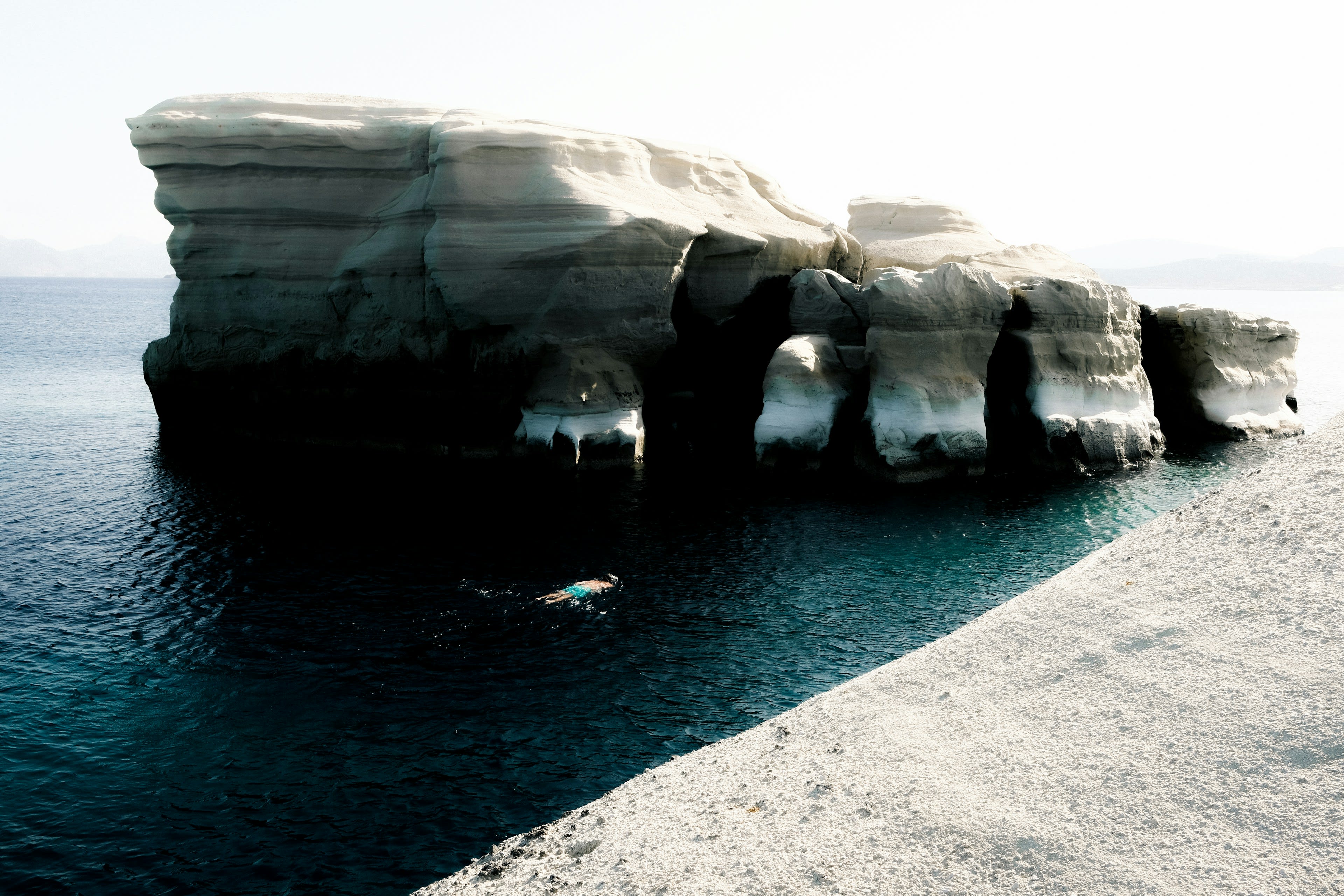 A person swimming in deep blue water beneath white rock cliffs