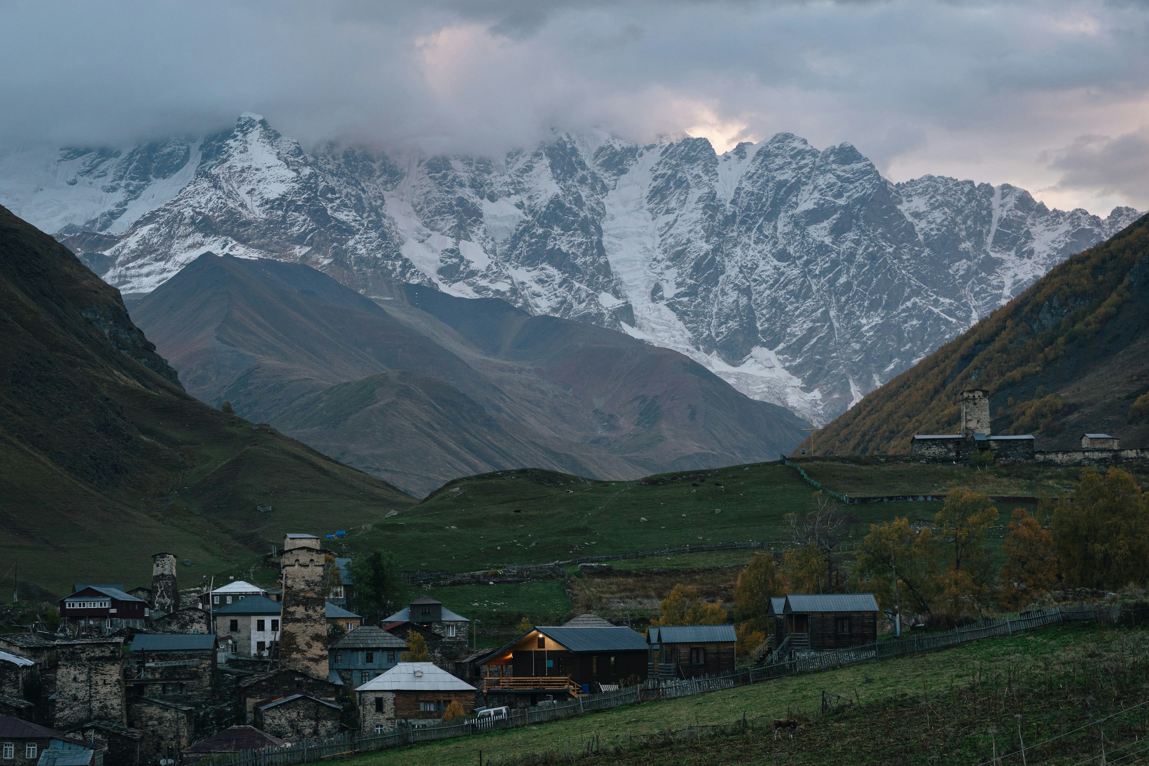 A village of stone towers in a green valley below a long snow-covered mountain wall