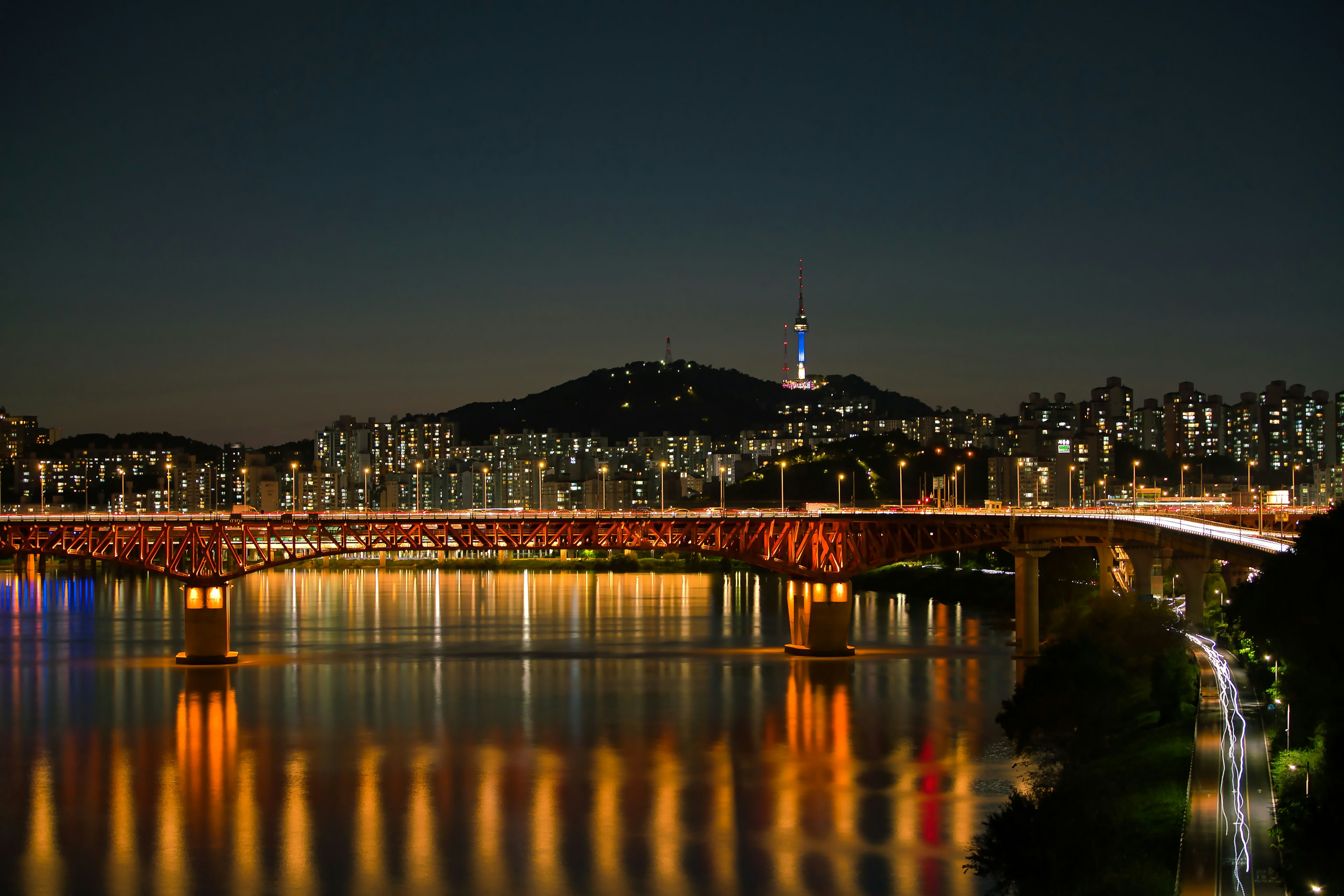 A lit bridge over a wide river with a city skyline and tower behind at night
