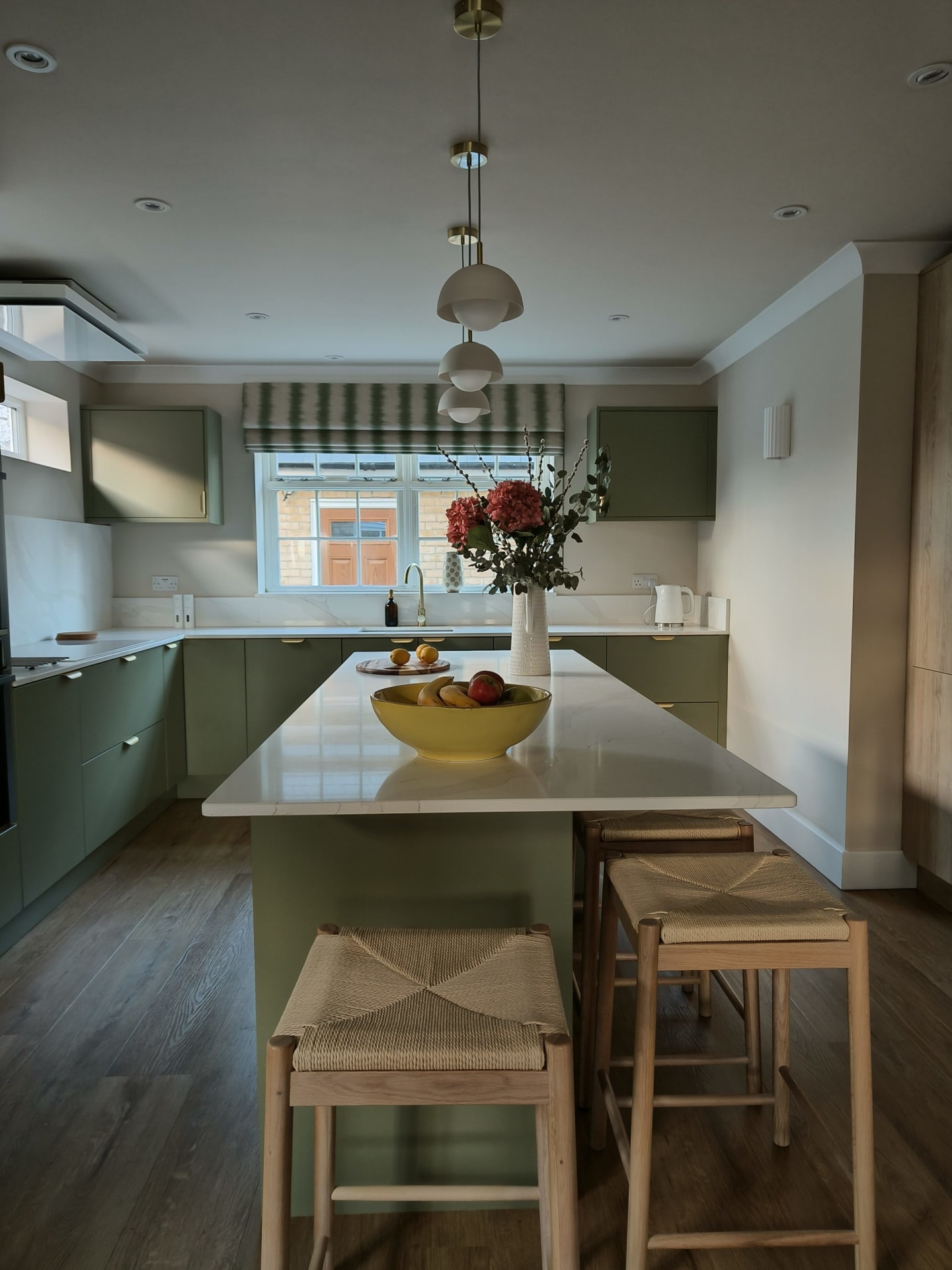 Sage green kitchen island with white quartz worktop, glass pendants and rattan stools