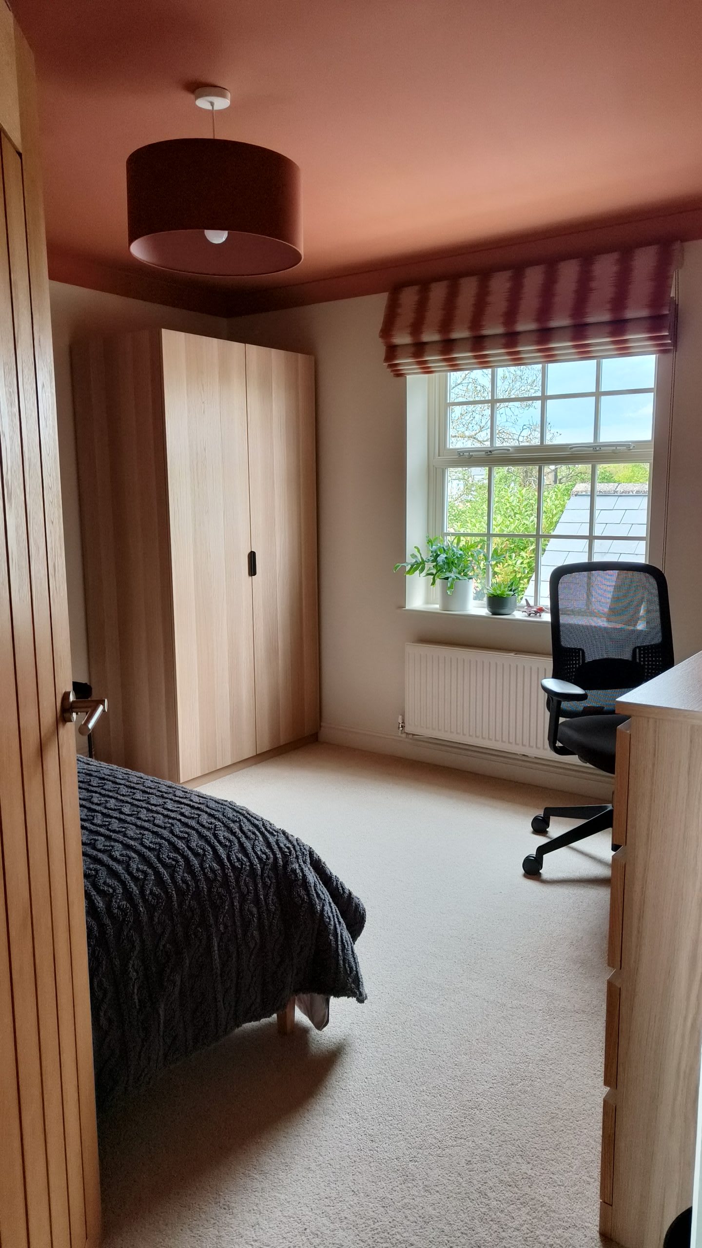 Bedroom corner with oak wardrobe, striped roman blind and a desk chair by the window