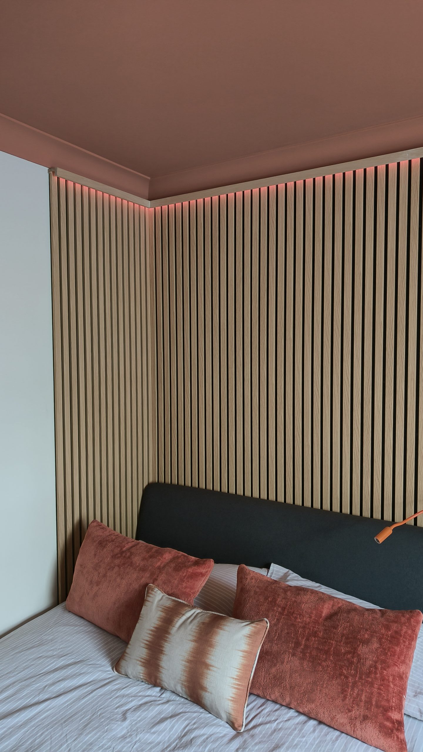 Backlit oak slat panelling behind a bed dressed with rust velvet cushions