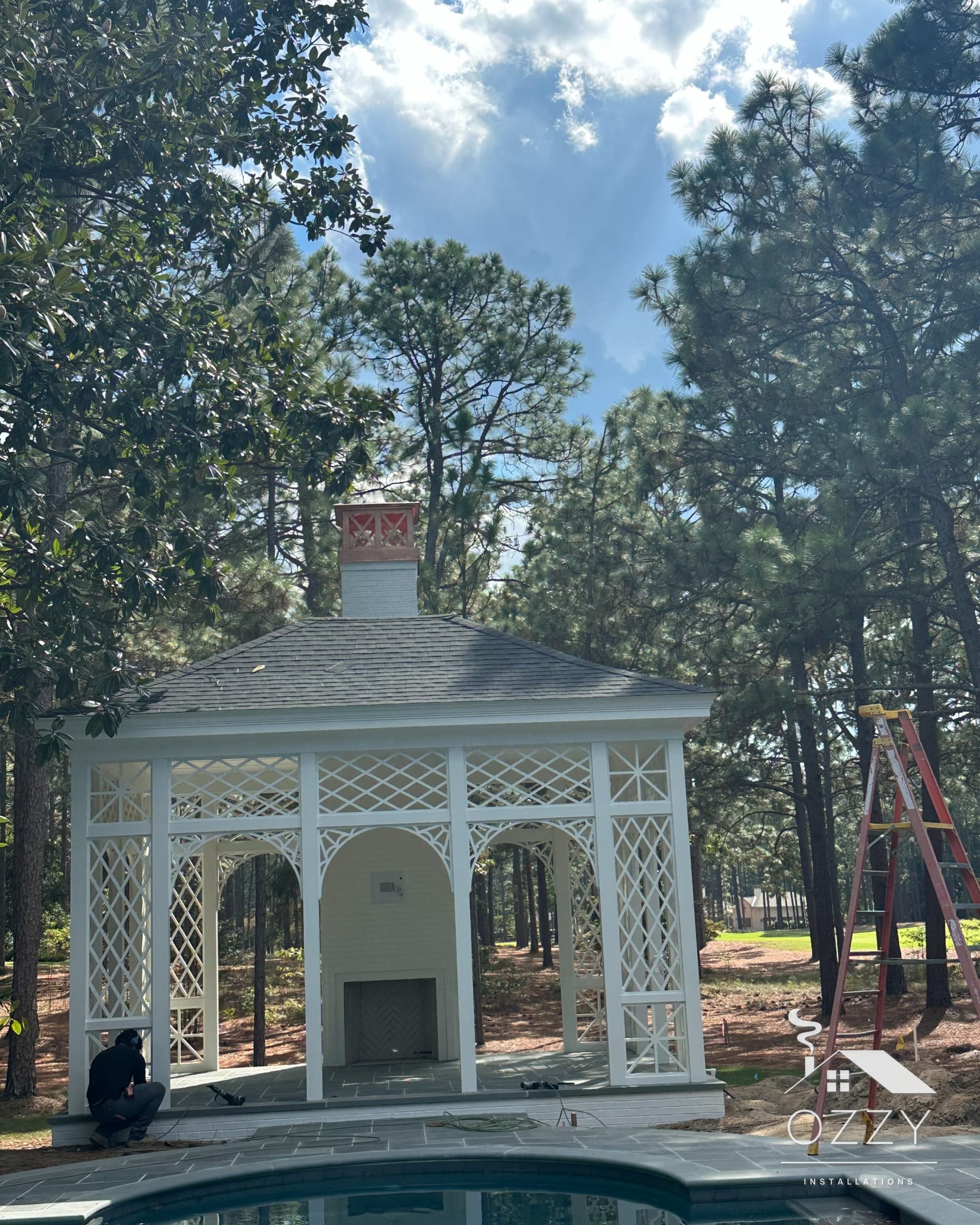 White lattice pool pavilion with a fireplace and chimney, seen across the pool