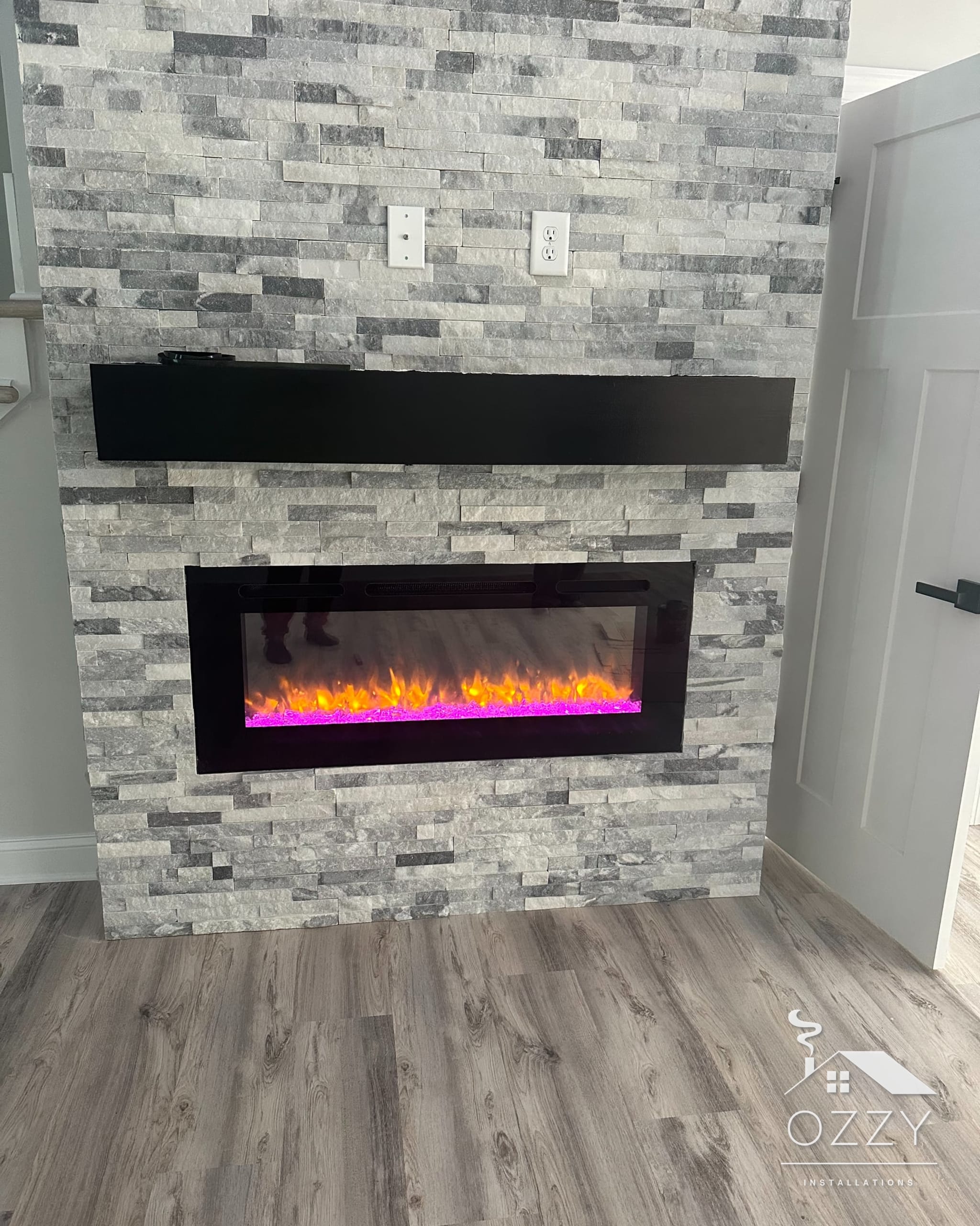 Linear electric fireplace with colored flames under a dark wood mantel in stacked stone