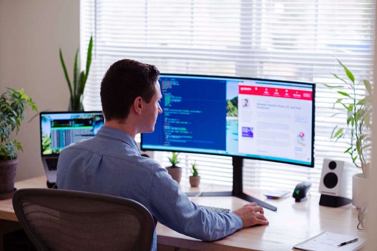 Freelance websites to hire python developers | Picture of a man in a blue collared shirt sitting at a desk and looking at a computer screen with code on one half of it and a website on the other half.