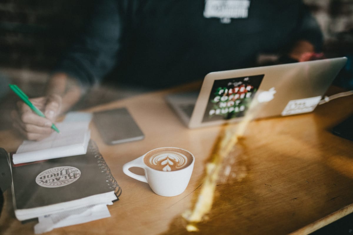 What can you develop with Java? | Picture shot through a window of a person working at a coffee shop with a laptop computer and a cup of coffee, writing in a notebook.