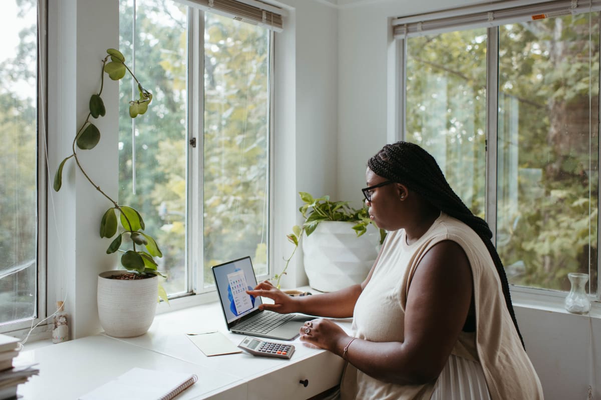 Woman in a cream colored tank top sitting at a white desk in a light-filled office. There are plants on the desk, and she is working at a laptop computer with a calculator to the left of the computer.