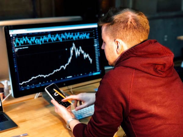 Shot from behind of a man in a red sweatshirt sitting at a computer that has a chart on it, while also scrolling on his phone.