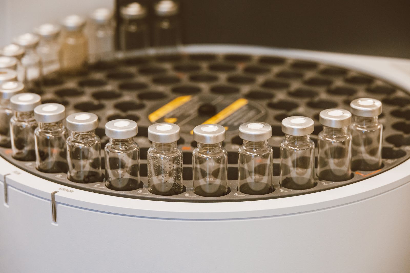 A row of clear glass vials with silver crimp caps standing in the round tray of a laboratory sample changer.