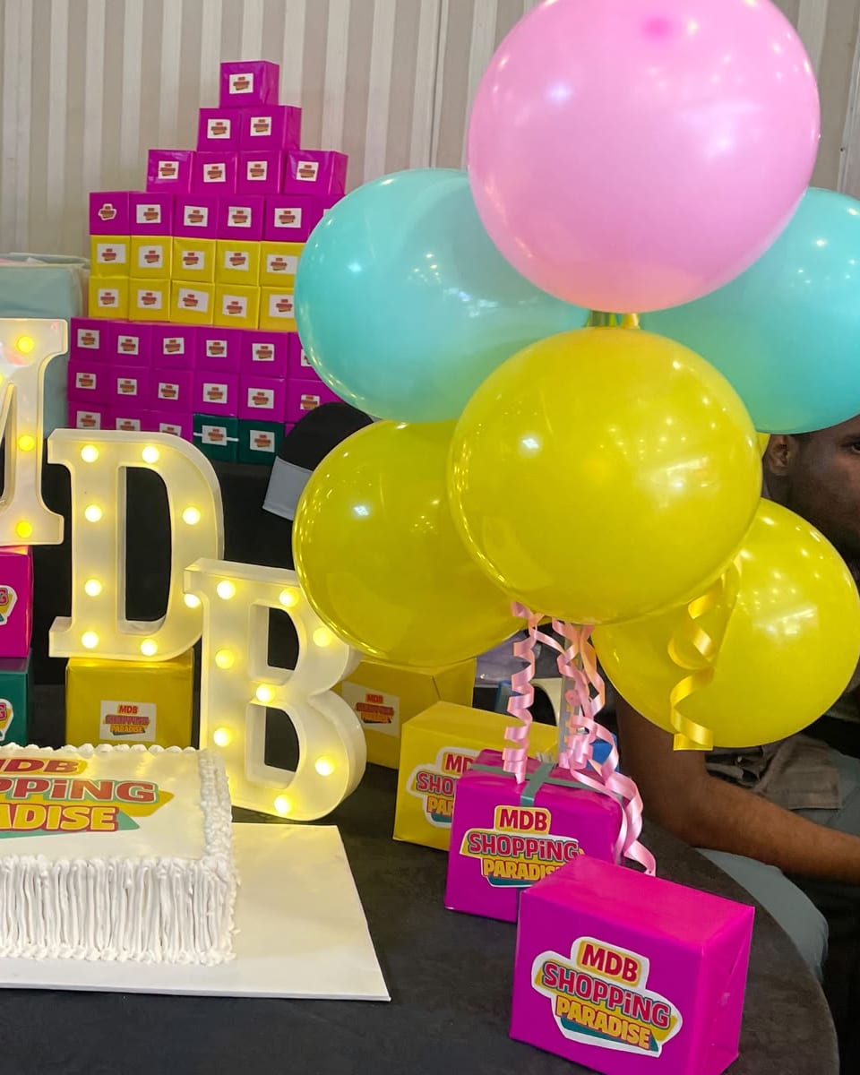 Pink, teal and yellow balloons, light-up MDB letters and a cake on a decorated table