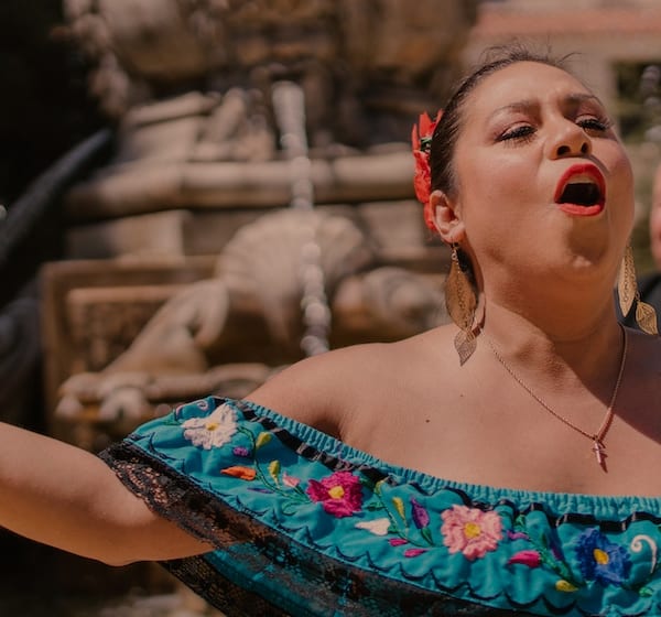 Traditional Hispanic artist singing with others playing instruments behind her.