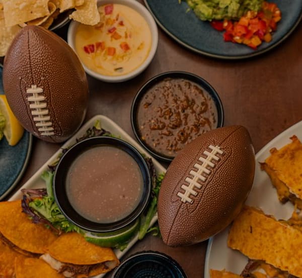 A table filled with Mexican appetizers like guacamole, quesadillas, tacos and queso paired with football decorations