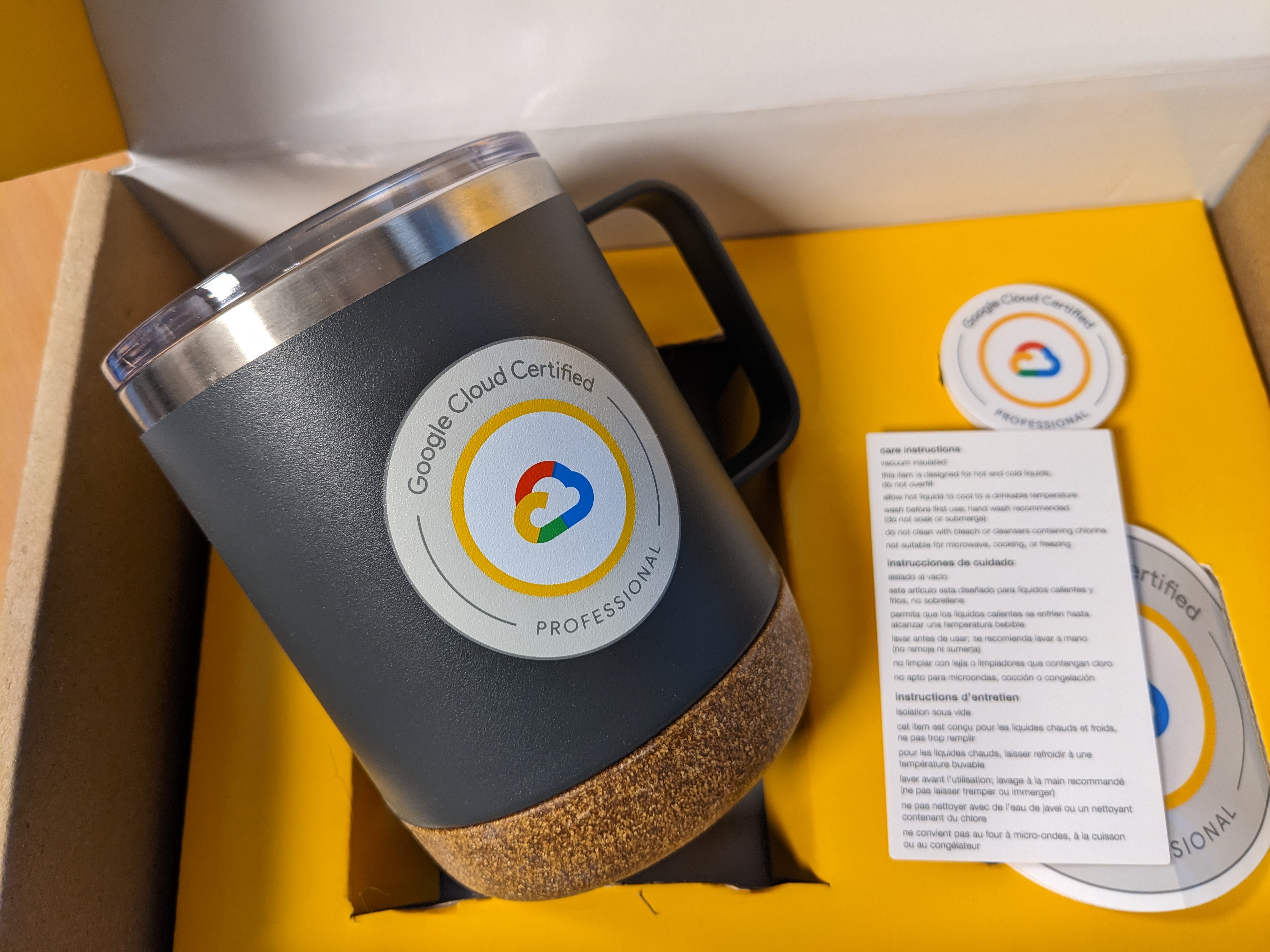 A dark grey Google Cloud mug with a cork bottom and a "Certified Professional" sticker, shown next to its care instruction card.