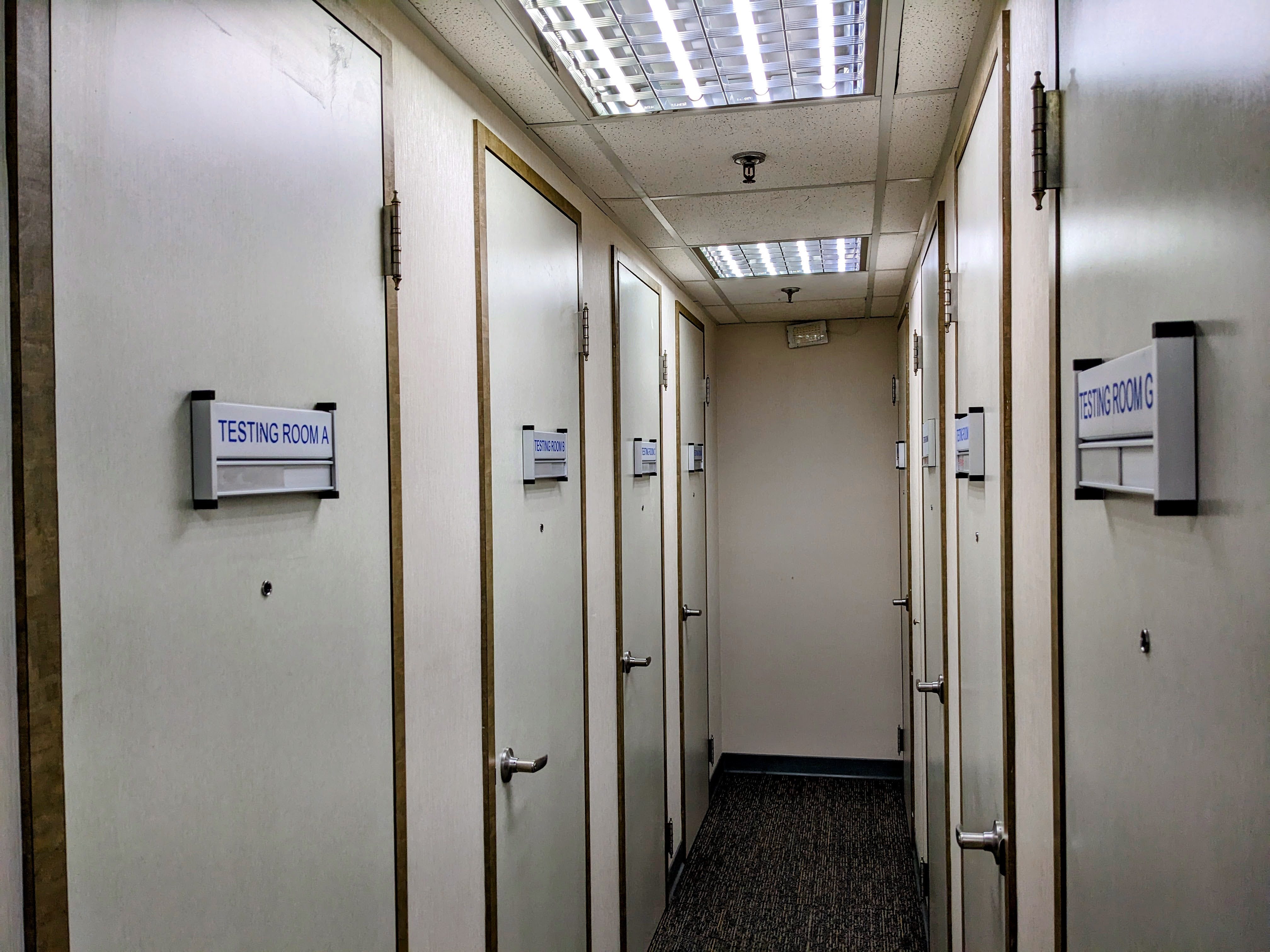 A narrow hallway inside a testing center, lined with several identical doors labeled with signs like "TESTING ROOM A" and "TESTING ROOM G."