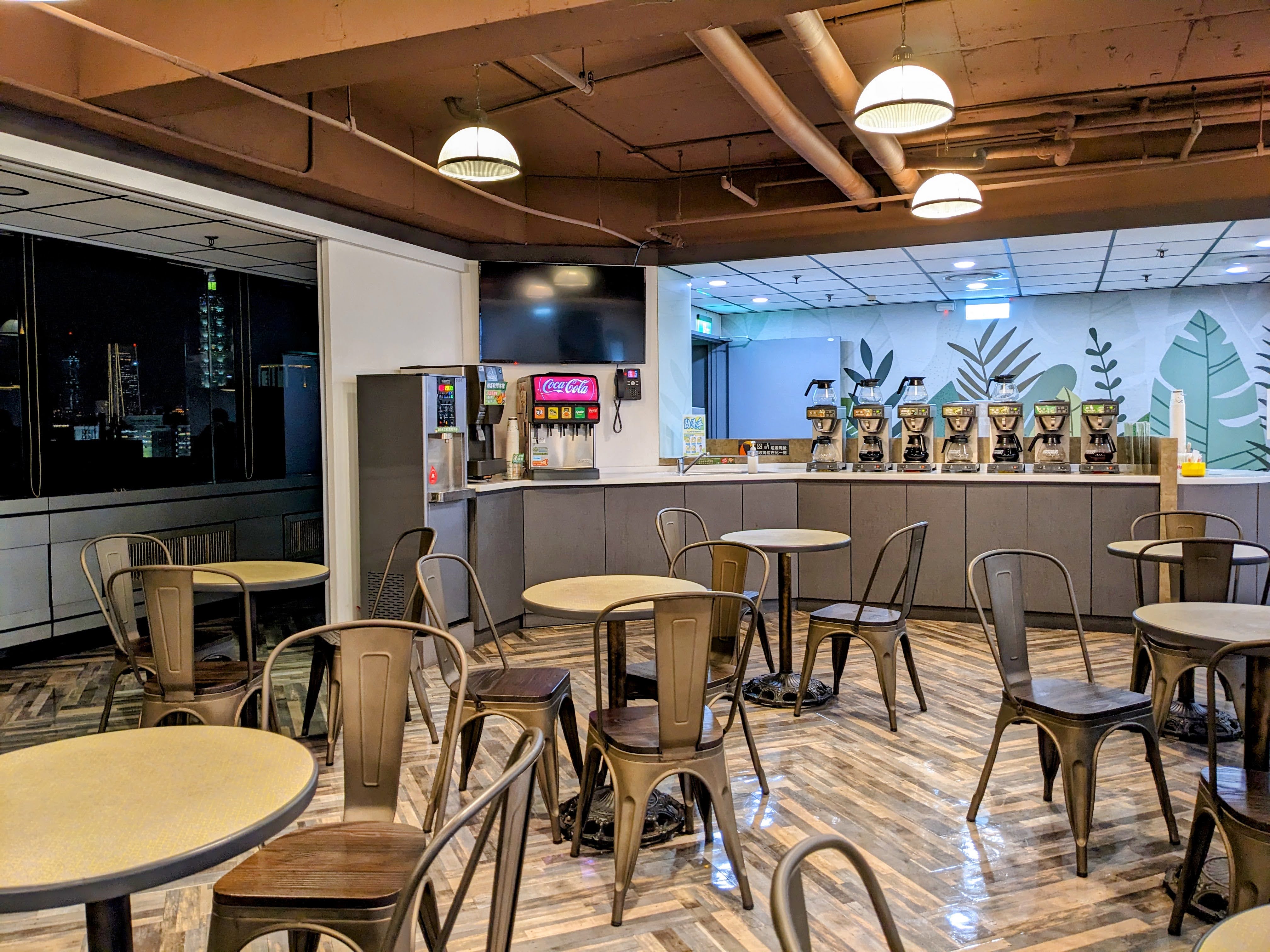 The modern lounge area of a testing center at dusk, with tables, chairs, a long counter with coffee and soda machines, and a view of the Taipei 101 skyscraper through a window.